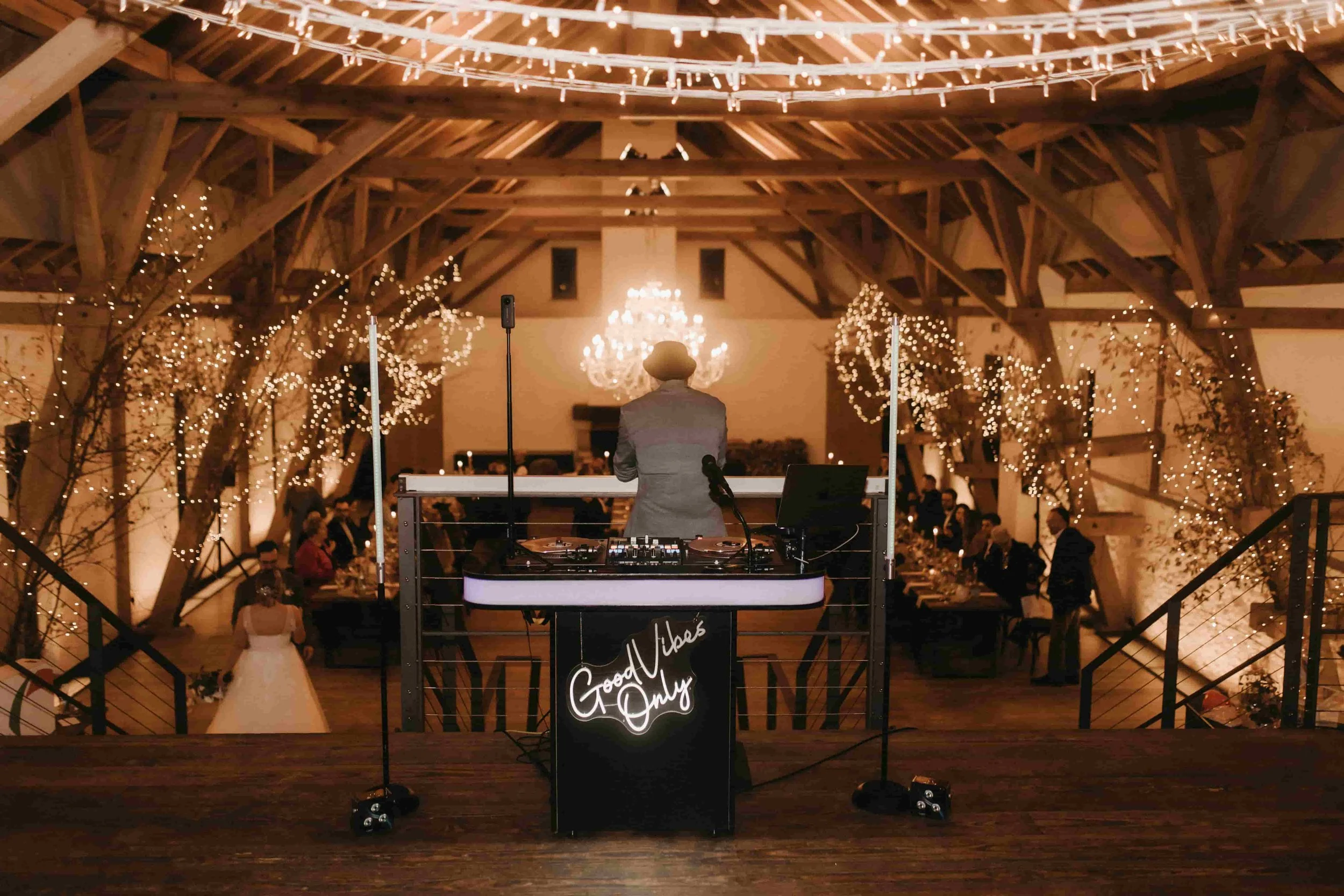 DJ an einem DJ-Pult auf einer Hochzeitsfeier mit Gästen im Hintergrund, festlich dekoriert mit Lichterketten, in einem Holzsaal mit Kronleuchtern