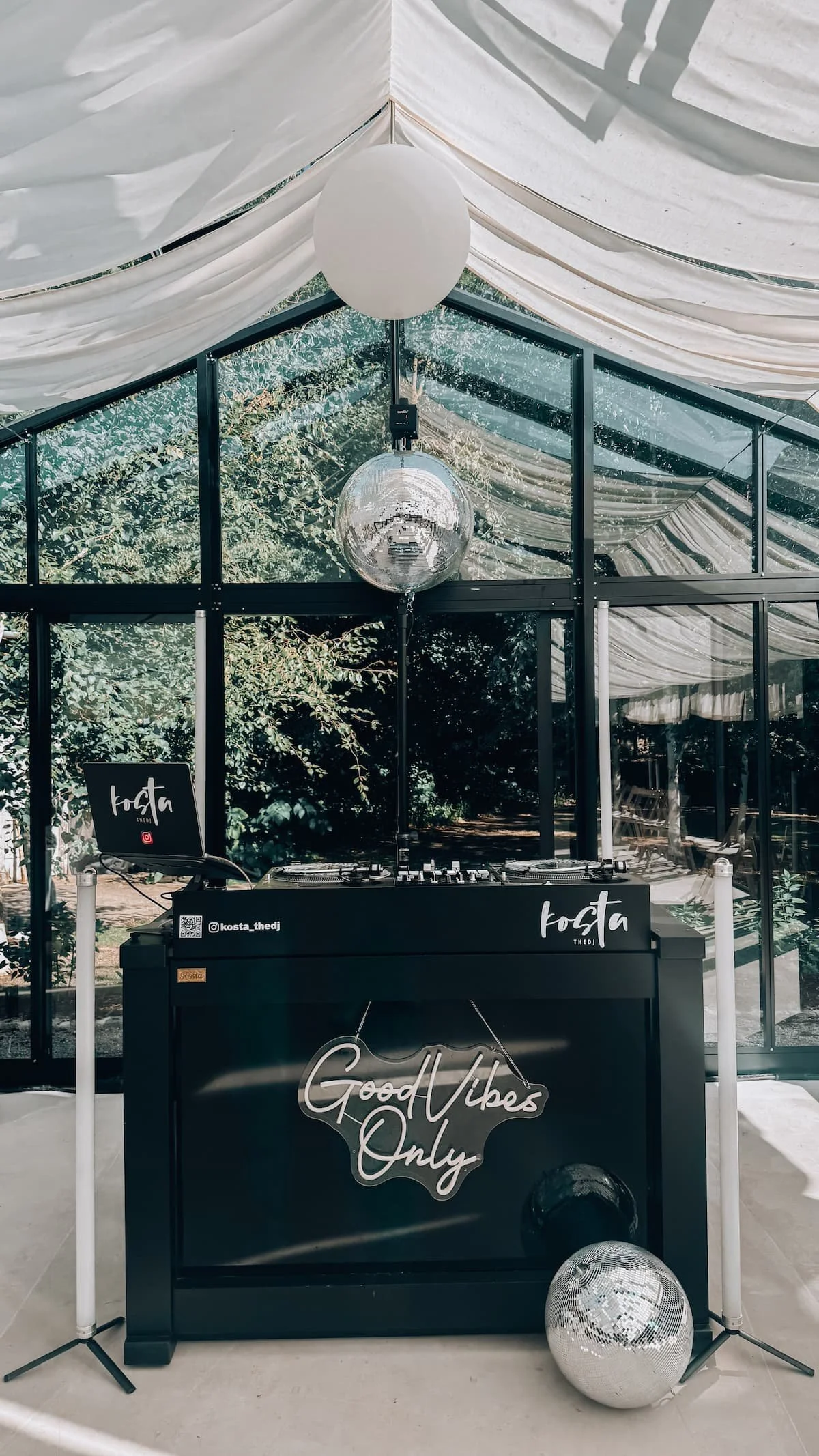 DJ-Station mit Laptop, Turntables und Discokugel in einem Zelt mit weißen Stoffbahnen, Dekoration mit Spiegelkugel, Pflanzen im Hintergrund, der Schriftzug 'Good Vibes Only' auf dem DJ-Tresen, eine weitere Discokugel auf dem Boden.
