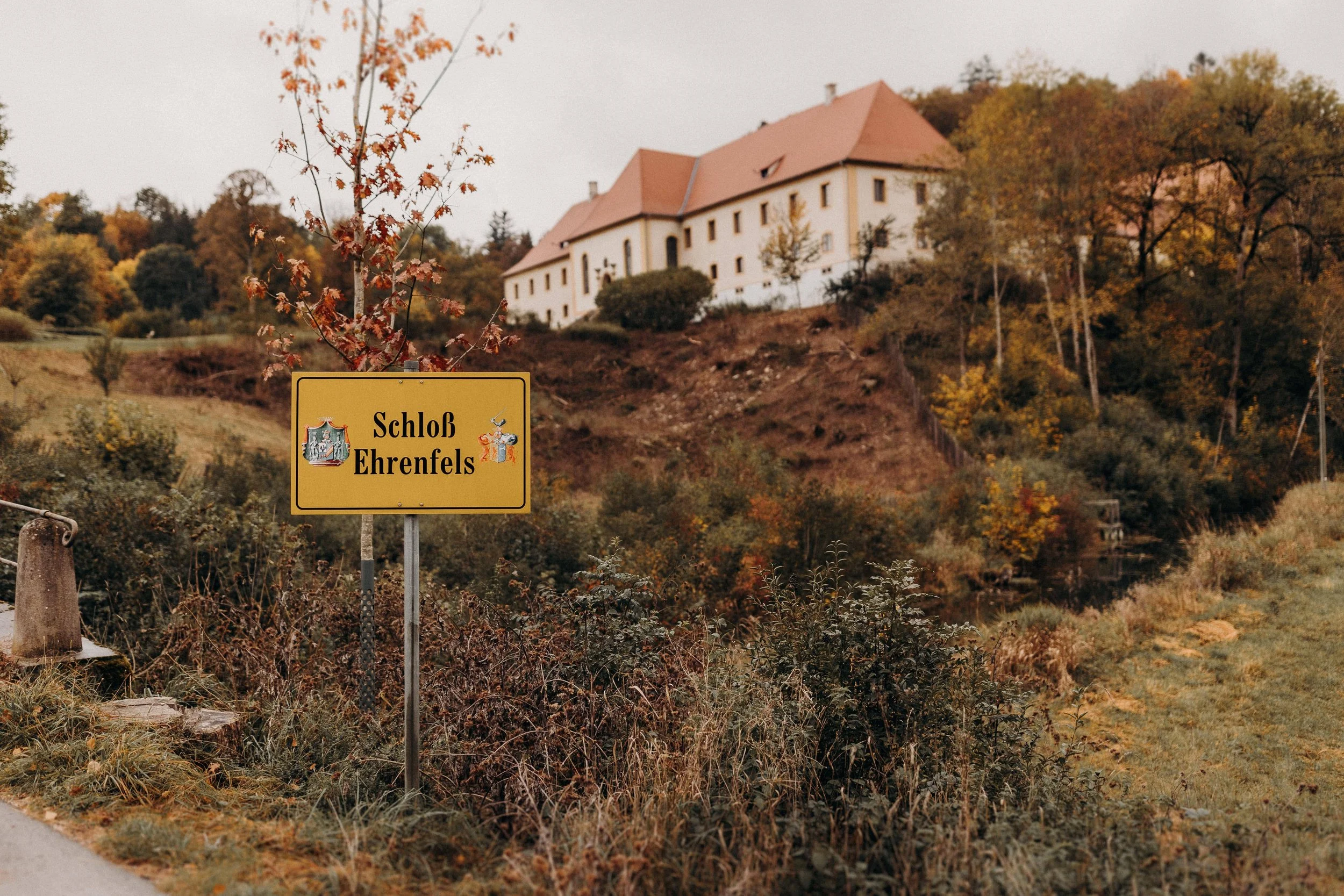 Schild mit Aufschrift 'Schloß Ehrenfels' vor einem Schloss auf einem Hügel, umgeben von Bäumen und Natur im Herbst.