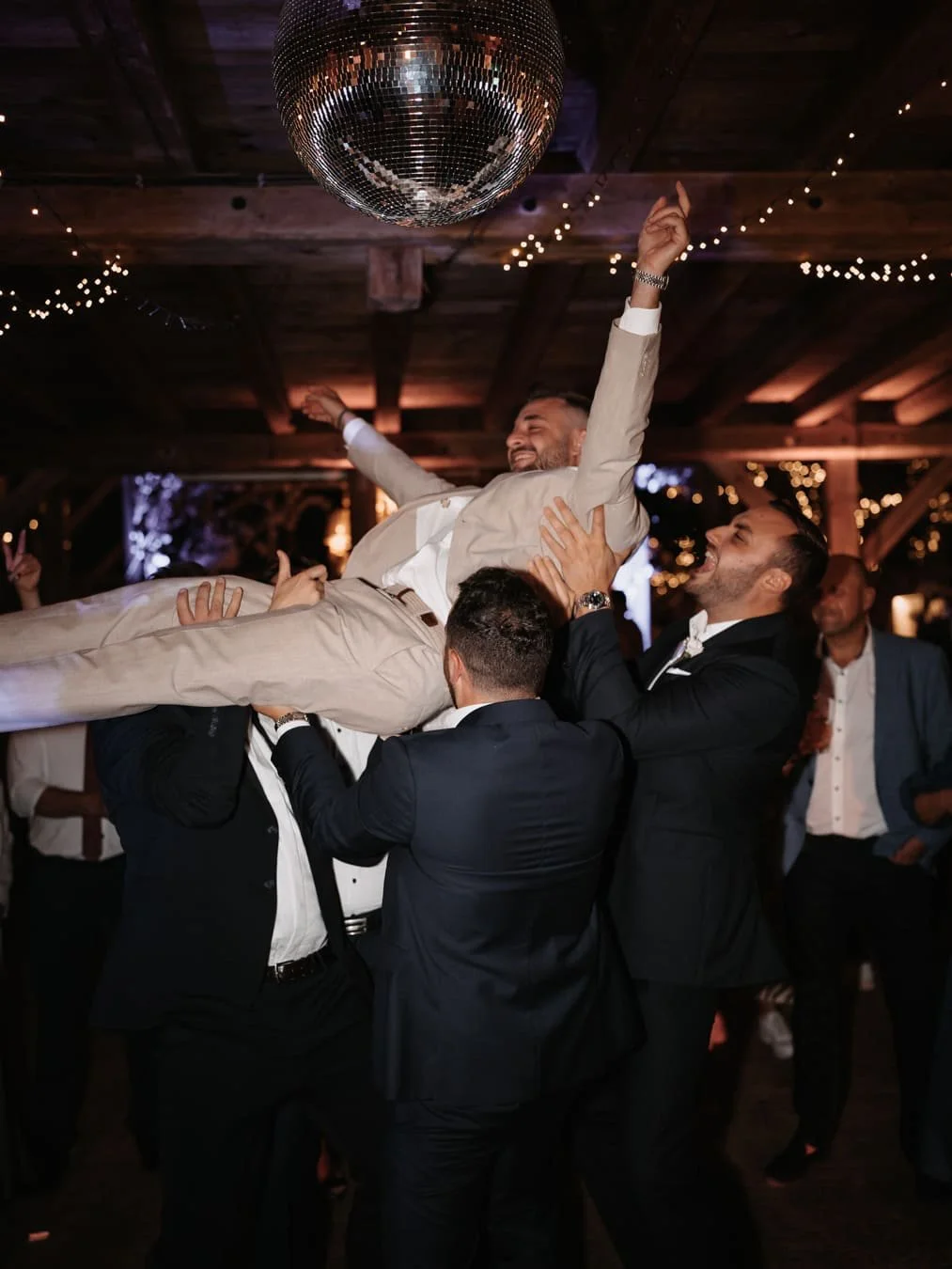 Männer feiern bei einer Hochzeit und heben den Bräutigam in die Luft, während er in einem festlichen Raum mit Lichterketten und Diskokugel ist.
