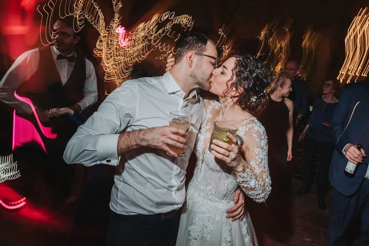 Ein Paar, das auf einer Hochzeit tanzt und sich küsst, während andere Gäste im Hintergrund feiern.