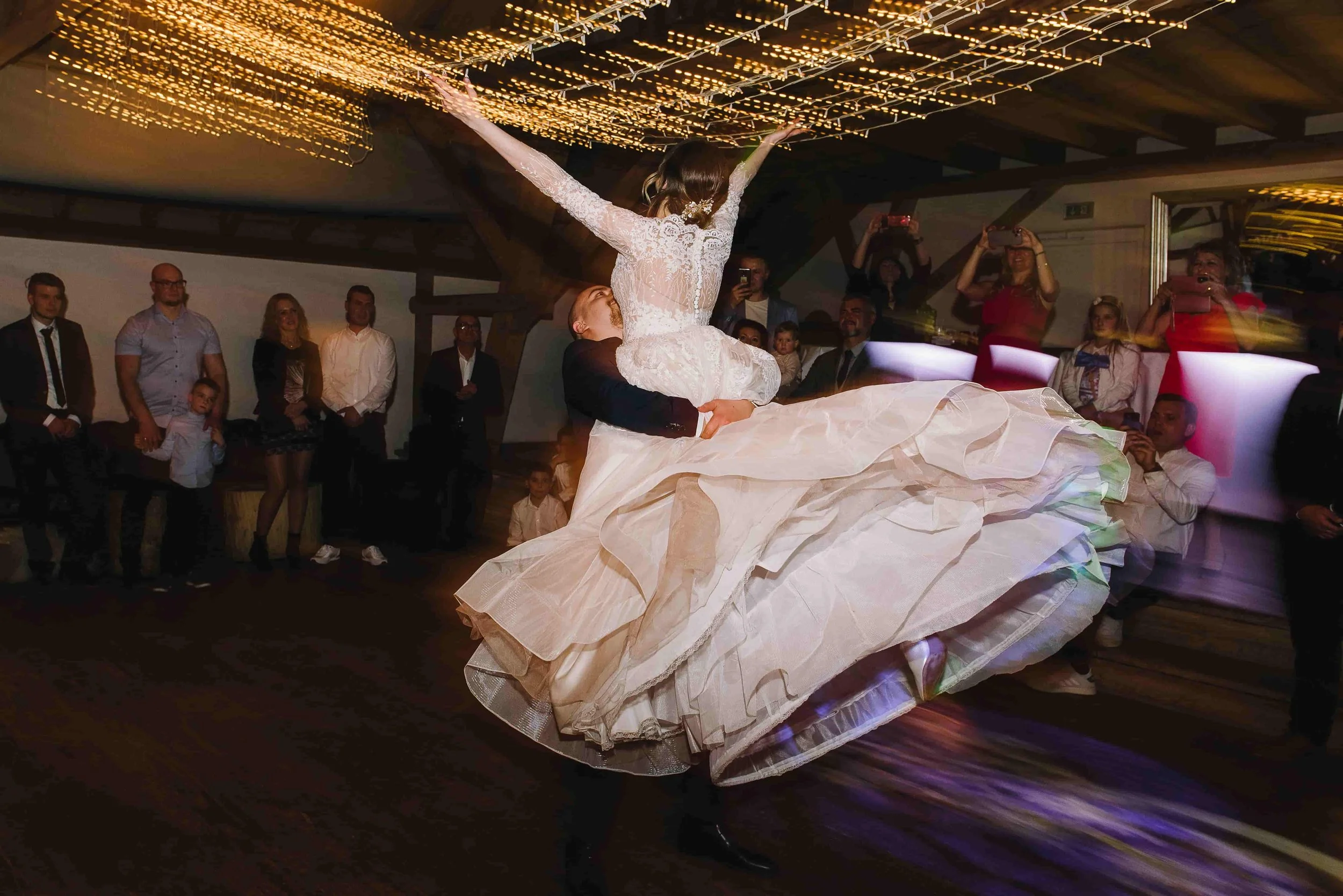 Braut wird bei der Hochzeit durch einen Mann auf einem Tanzboden hoch gehoben, um einen Tanz aufzuführen. Im Hintergrund beobachten Gäste die Szene und machen Fotos.