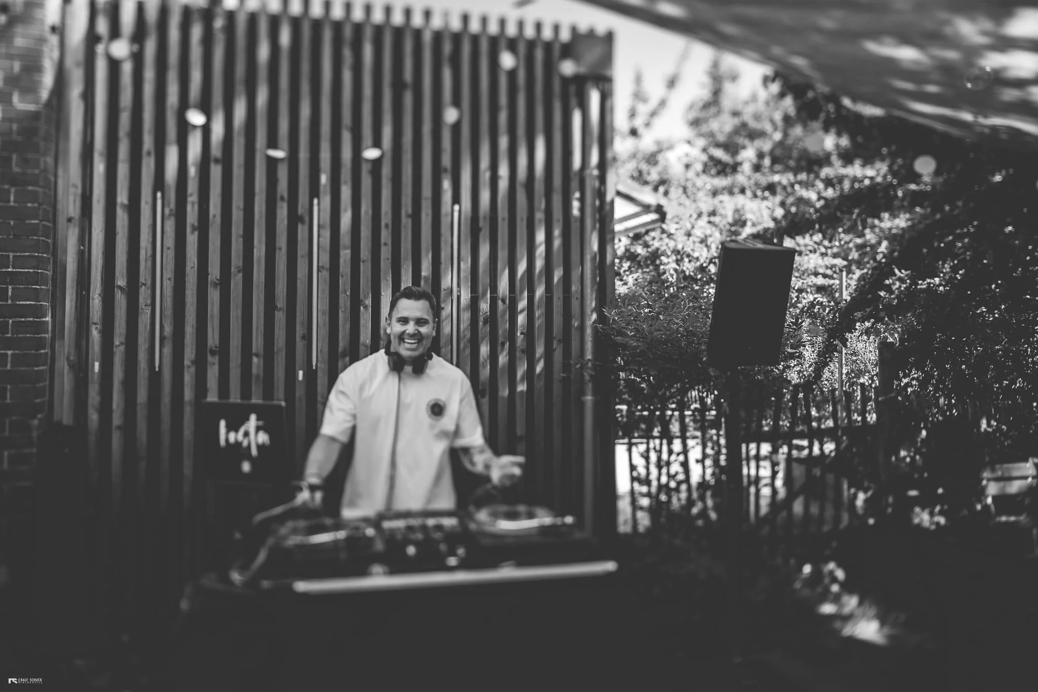 Eine lachende Person mit DJ-Equipment bei einer Outdoor-Veranstaltung, im Hintergrund eine Holzlattung und Bäume.