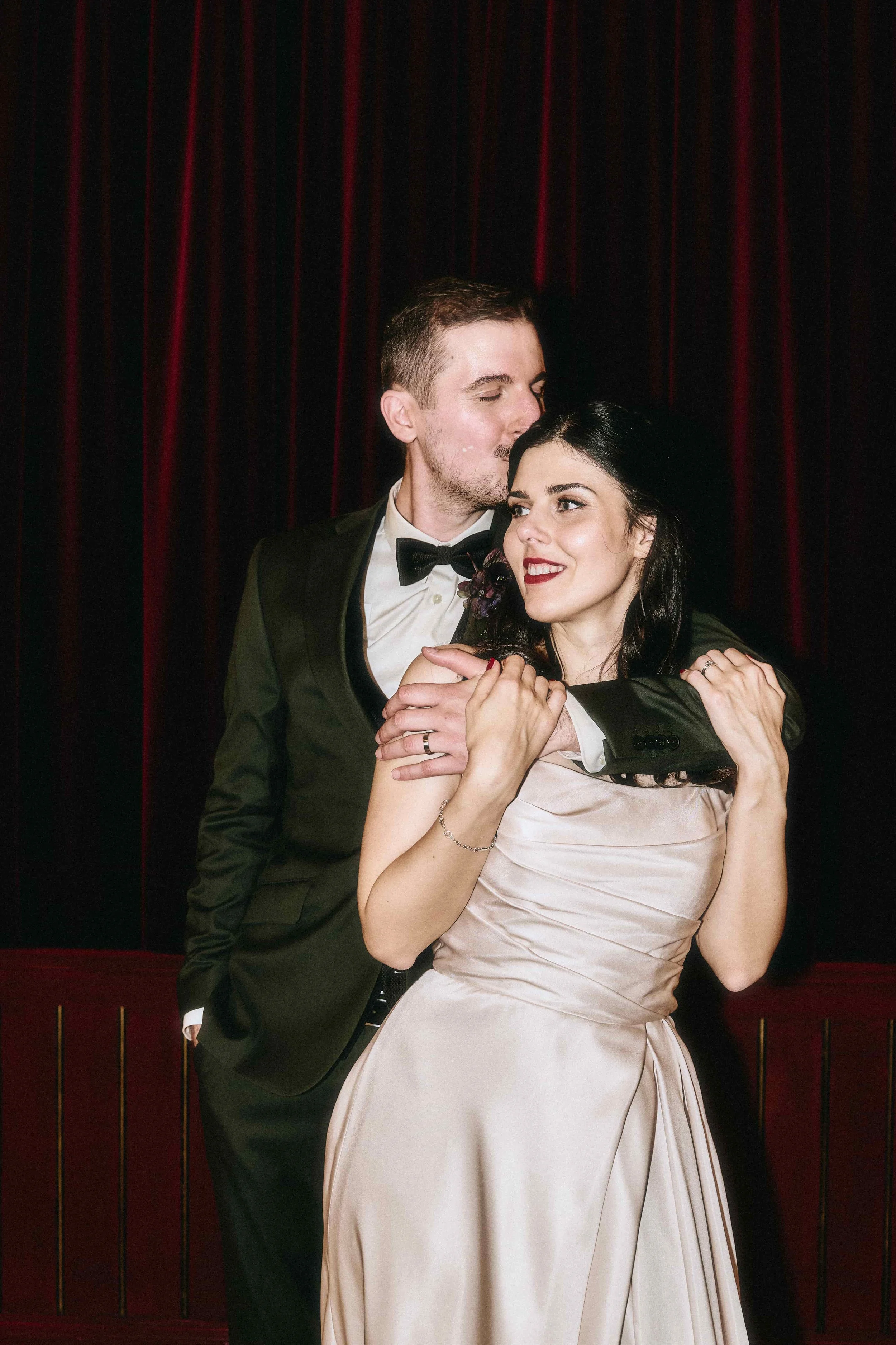 Ein Paar in formeller Kleidung, die sich umarmen und sich küssen, vor einem schwarzen Vorhang mit roten Streifen.