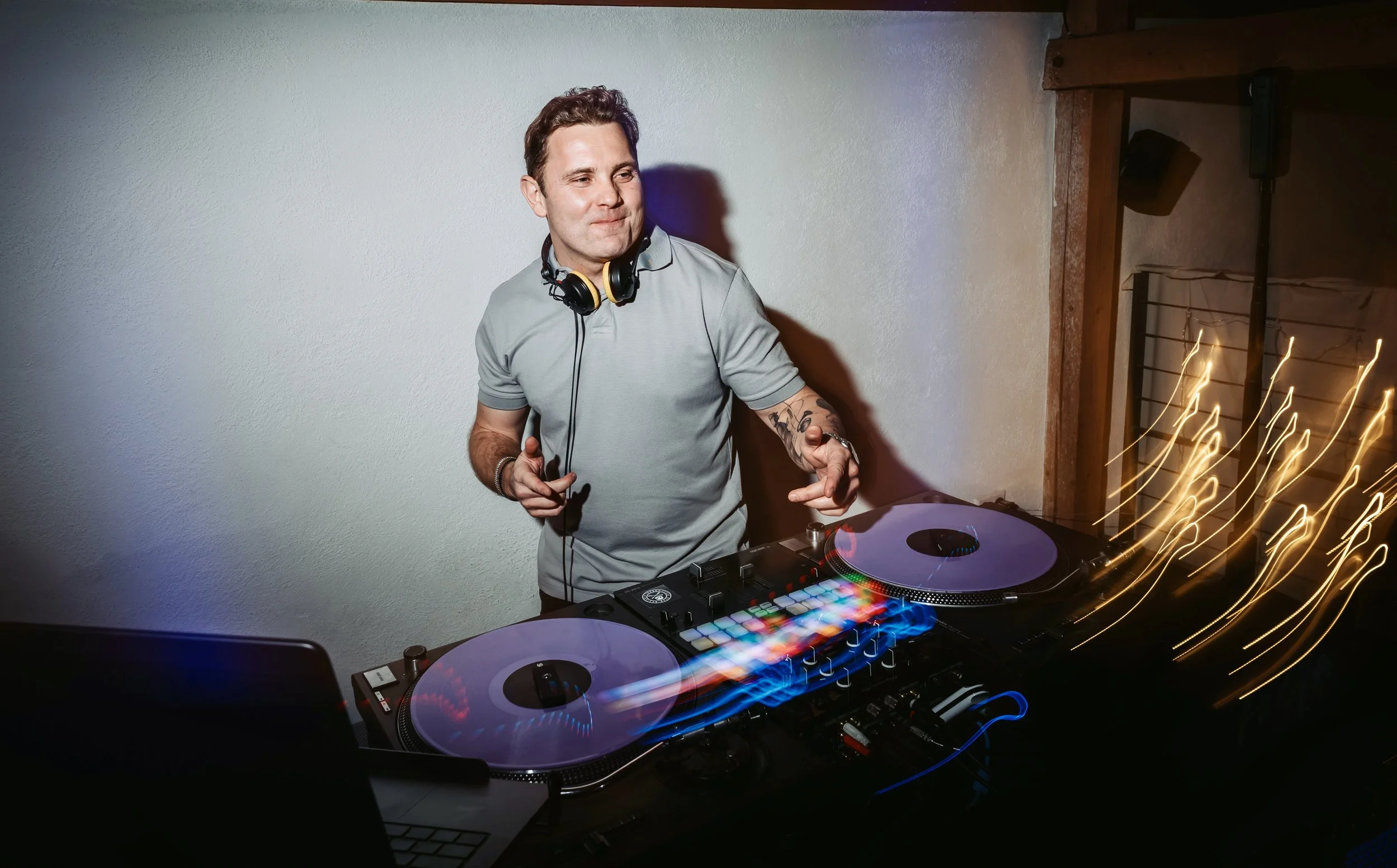 Ein junger Mann, der als DJ arbeitet, steht an einem DJ-Setup mit Turntables und einem Mixer, trägt Kopfhörer um den Hals und hat Tattoos am linken Arm, während er in einem dunklen Raum auflegt.