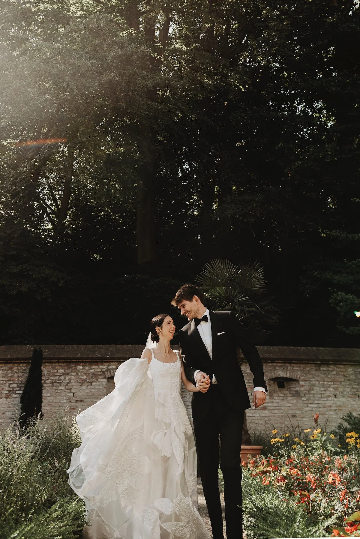 Ein Brautpaar beim Spazieren im Garten, die Frau trägt ein weißes Hochzeitskleid und der Mann einen schwarzen Anzug, sie halten Händchen und lachen.