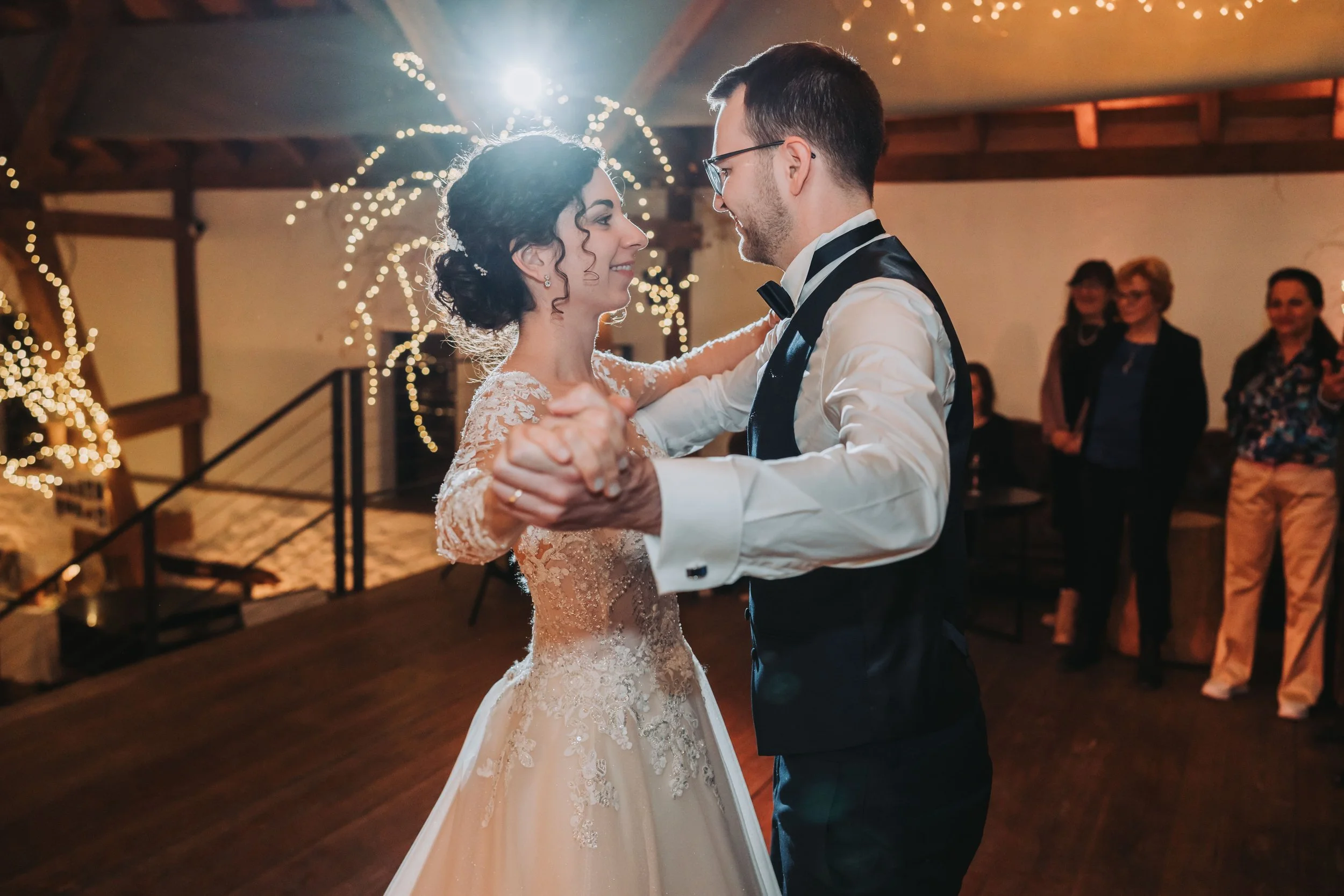 Ein Brautpaar tanzt auf einer Hochzeit, im Hintergrund sind Gäste und festliche Dekoration mit Lichterketten.