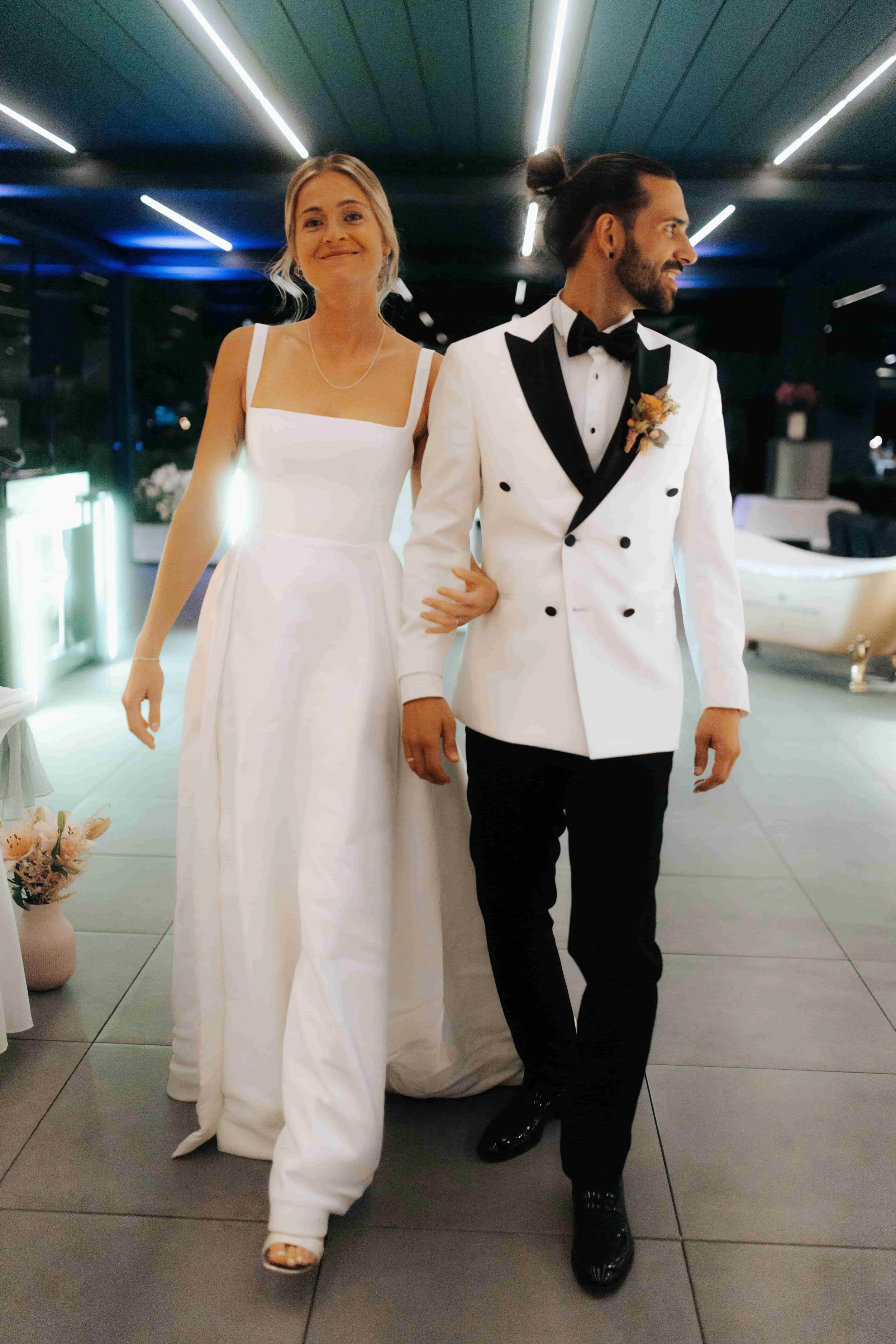 Ein Brautpaar bei ihrer Hochzeit, die Frau trägt ein weißes Brautkleid und der Mann einen weißen Smoking mit schwarzen Details, sie gehen durch einen Raum mit moderner Beleuchtung.