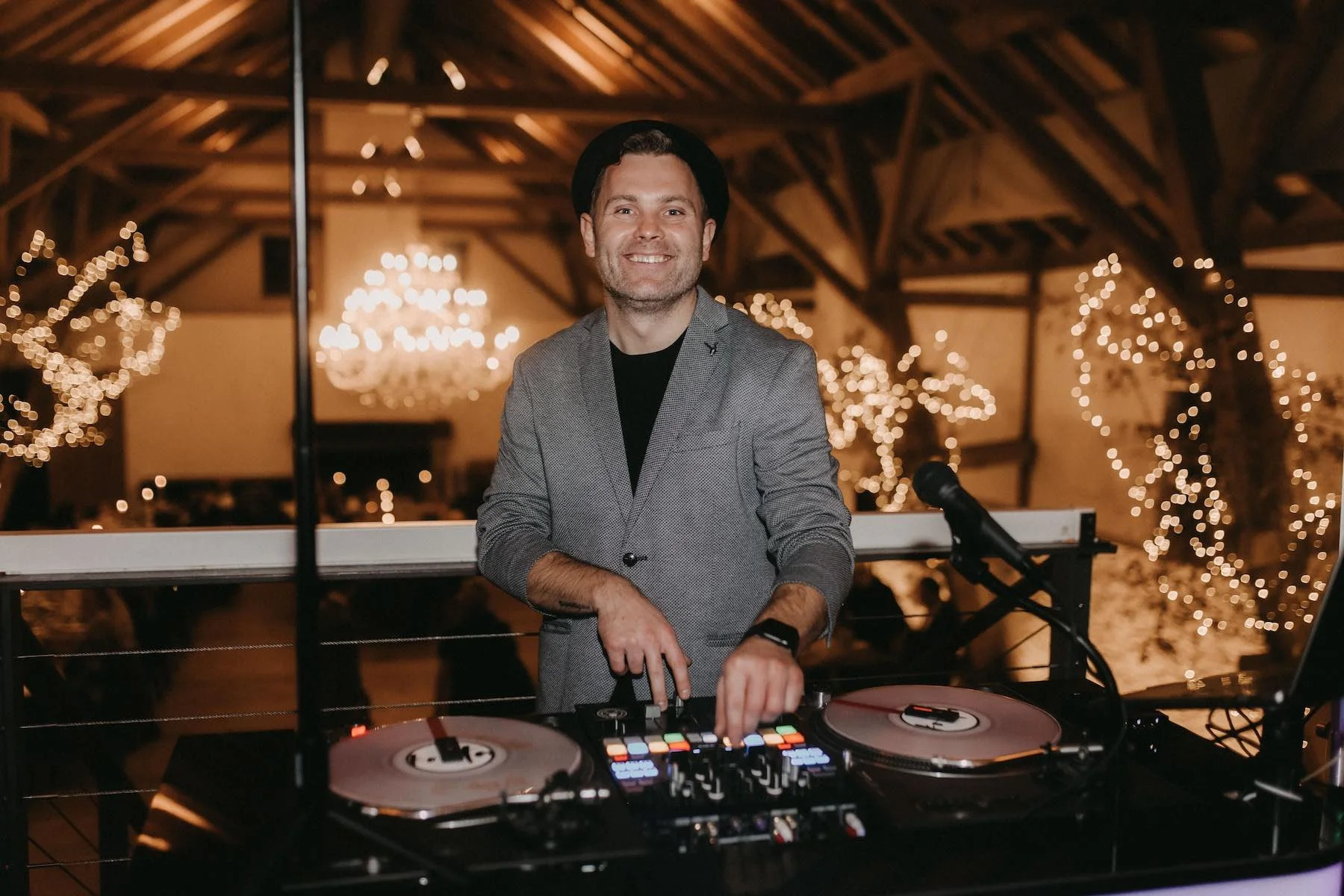 Mann als DJ hinter einem Mischpult, lachend, in einem Raum mit geschnörkelten Lichterketten an den Wänden, im Hintergrund Kronleuchter und Holzdecke.
