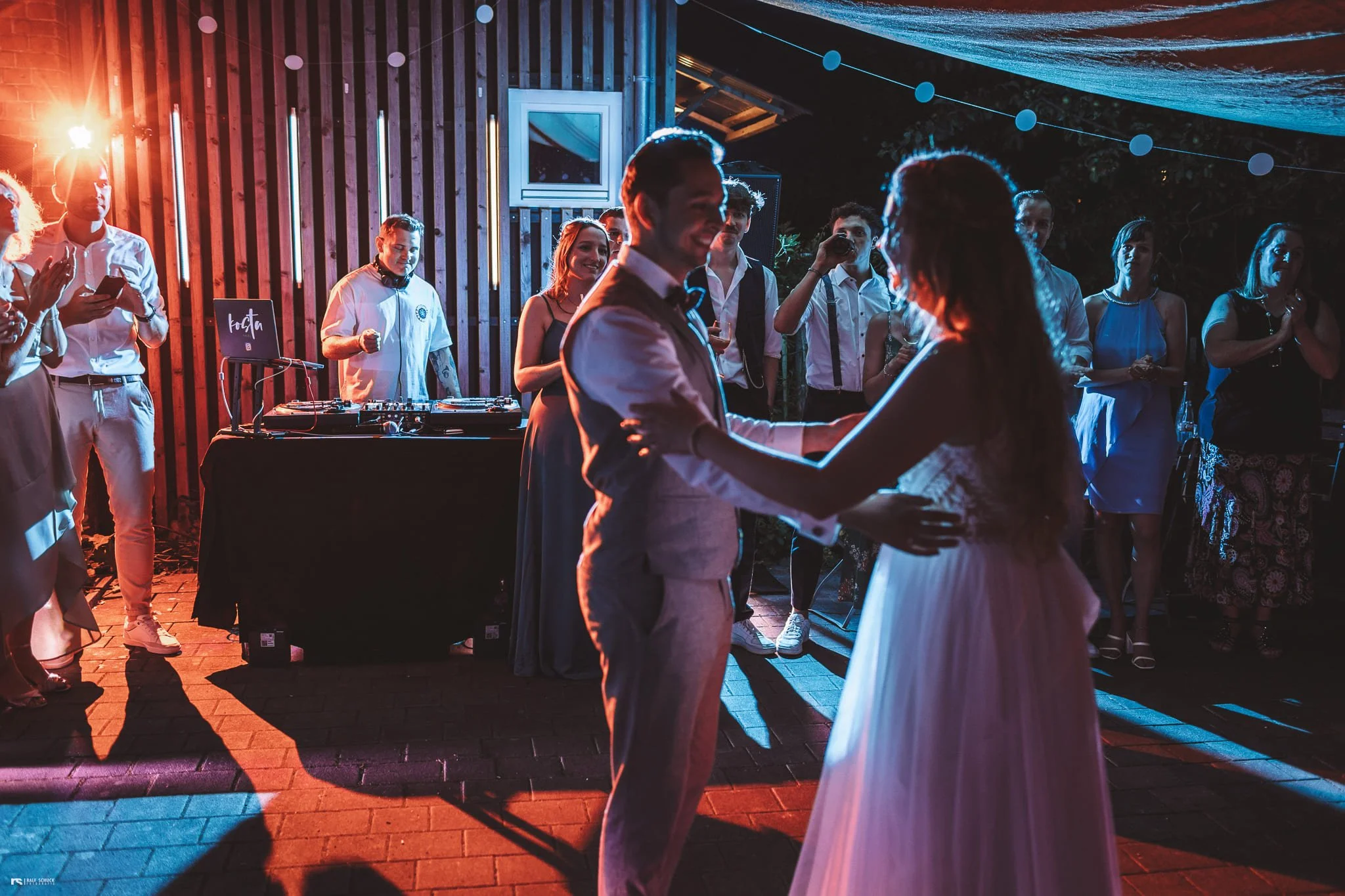 Ein Paar tanzt bei einer Hochzeit im Freien, im Hintergrund stehen Gäste und ein DJ mit Musikdj-Equipment, bei Nacht mit bunten Lichtern.