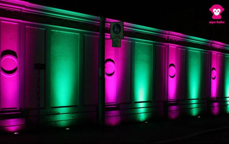 Gebäude mit pink und grünen LED-Lichtern beleuchtet, mit Mülleimer und Zonen-Schild davor, nachts aufgenommen.