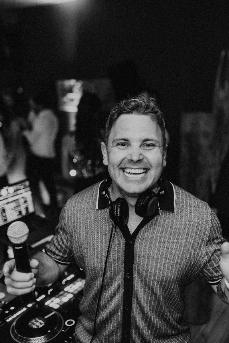 Ein glücklicher DJ mit einem Mikrofon in der rechten Hand, umgeben von Menschen in einer Party- oder Club-Umgebung, schwarz-weiß Foto.