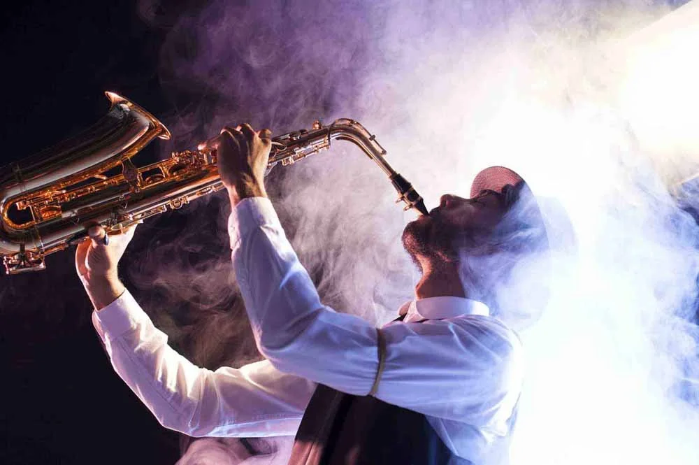 Mann spielt Saxophon in einer dunklen, mit Rauch und hellem Licht dramatisch beleuchteten Umgebung.