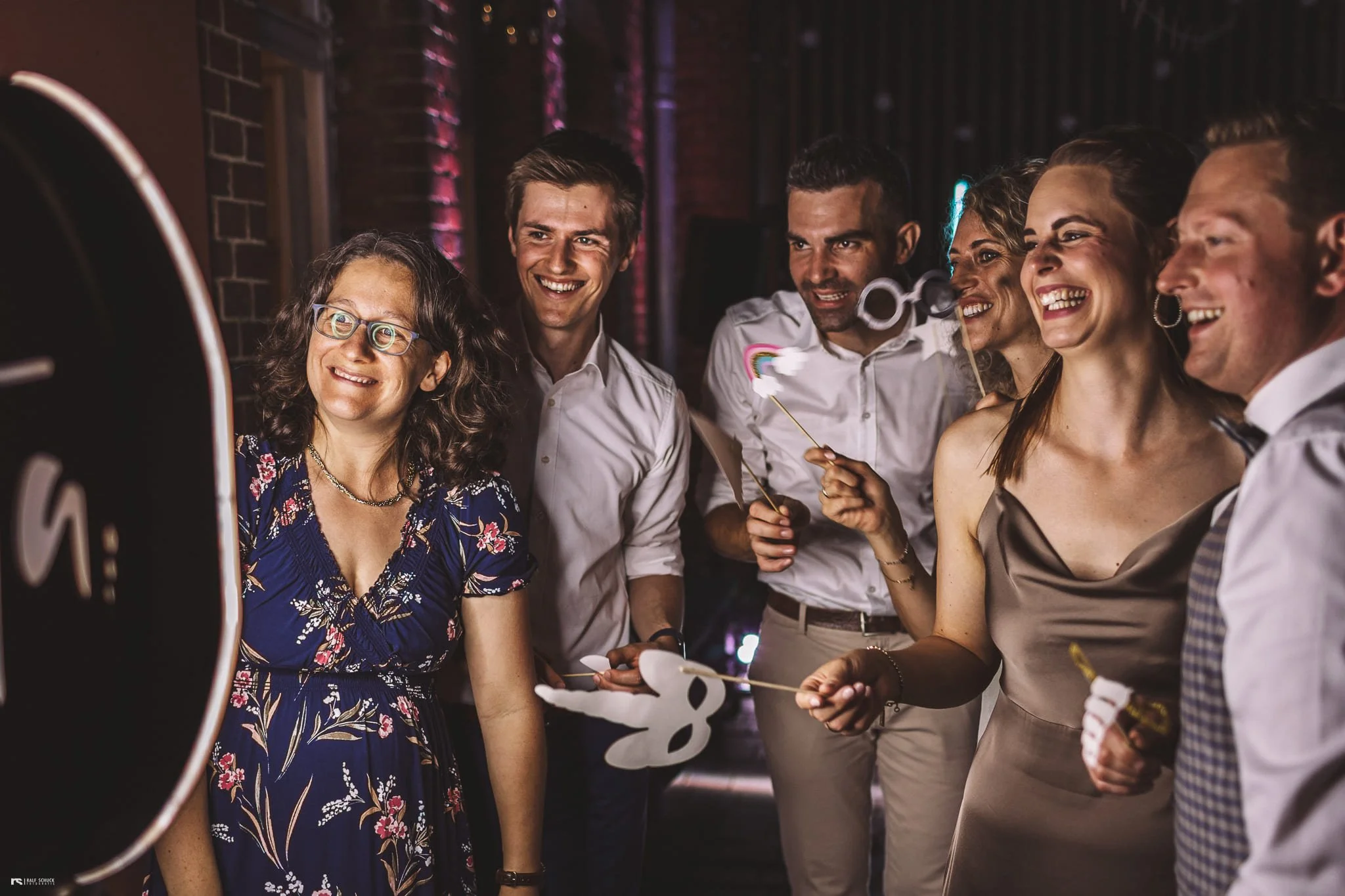 Gruppe von sechs Menschen bei einer Feier, die zusammen ein Foto machen, einige halten Himmel-und-Herz-Werden-Requisiten. Sie lachen und freuen sich in einem festlichen, gut beleuchteten Raum.