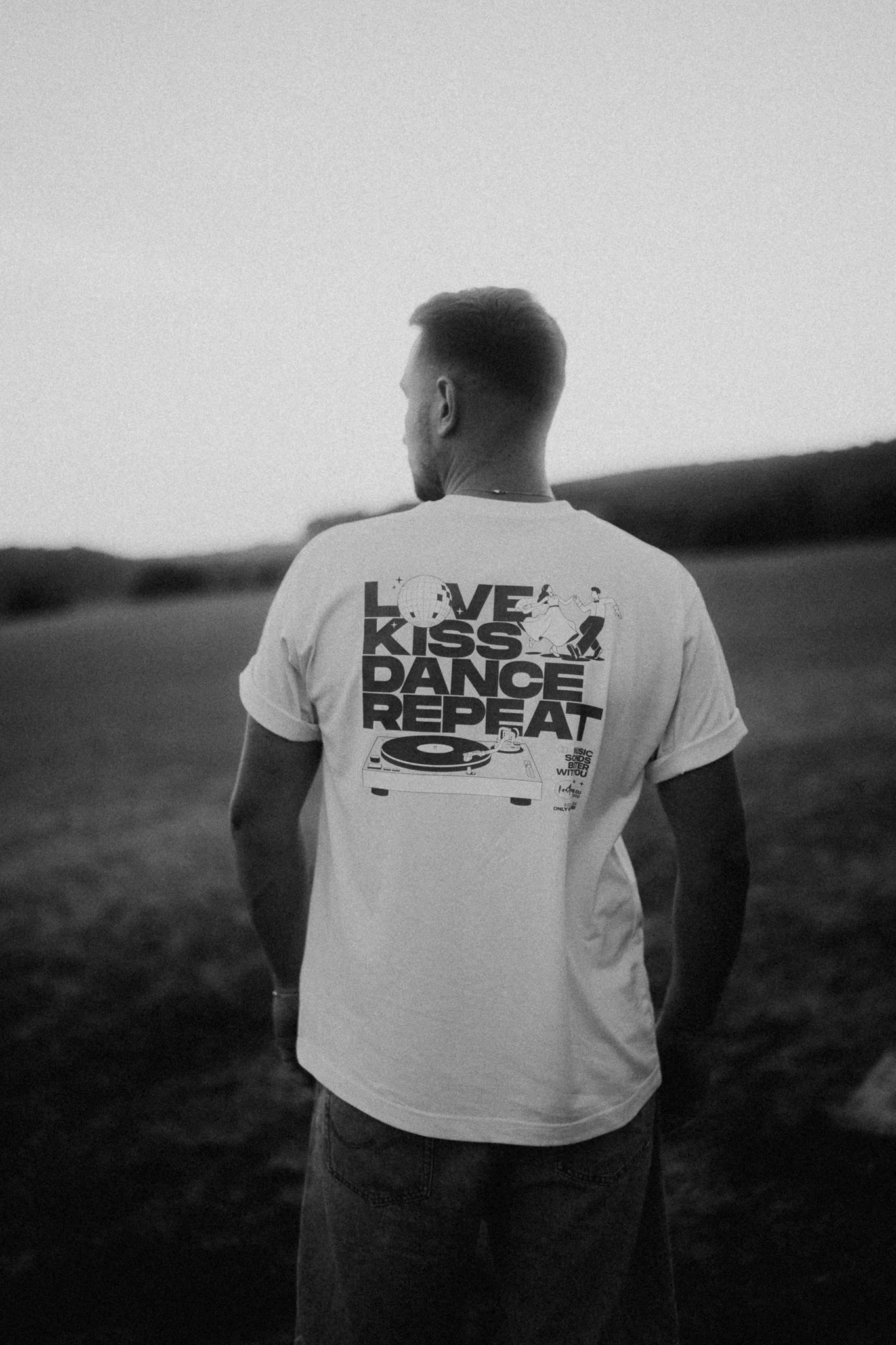 Ein junger Mann in einem weißen T-Shirt mit Musikmotiv, der nach hinten blickt, steht in einer ländlichen Umgebung im Abendlicht.