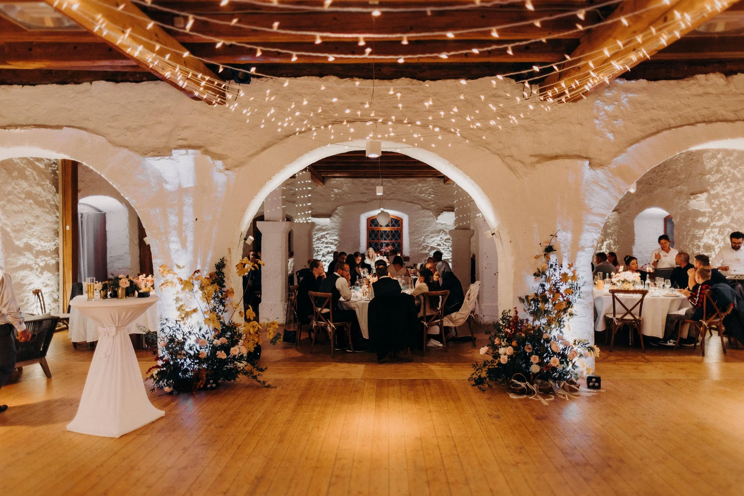 Innenraum einer festlich gedeckten Veranstaltung mit mehreren Tischen, weissen Wänden, Holzboden, Dekoration mit Lichterketten und Blumen, Gäste sitzen und unterhalten sich.