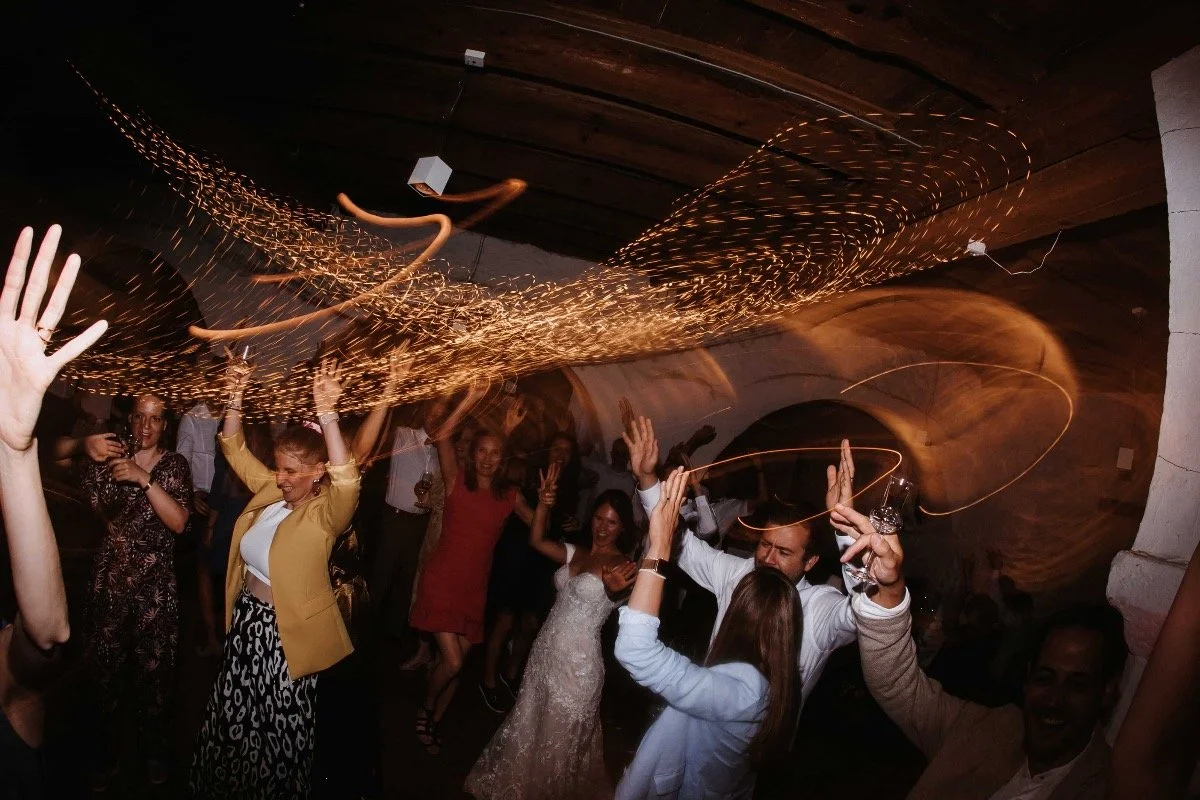 Menschen feiern bei einer Party, einige tanzen und heben die Hände, Lichterketten und ein Weinglas sind sichtbar.