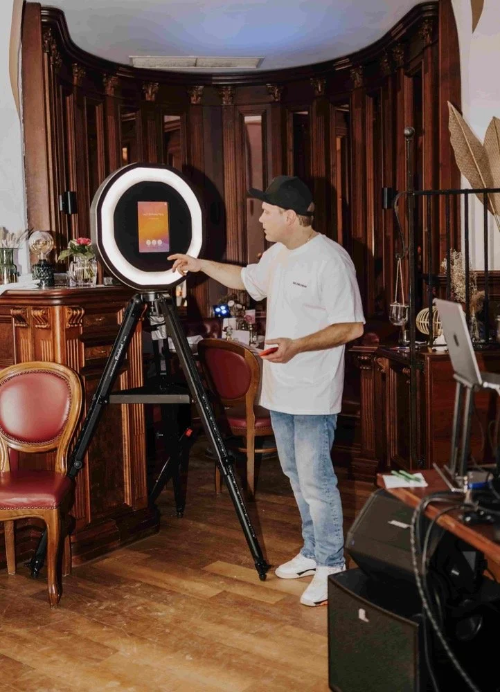 Ein Mann in einem weißen T-Shirt und einer schwarzen Cap benutzt eine Kamera mit einem Ringlicht in einem Raum mit dunklem Holzinterieur und roten Stühlen.