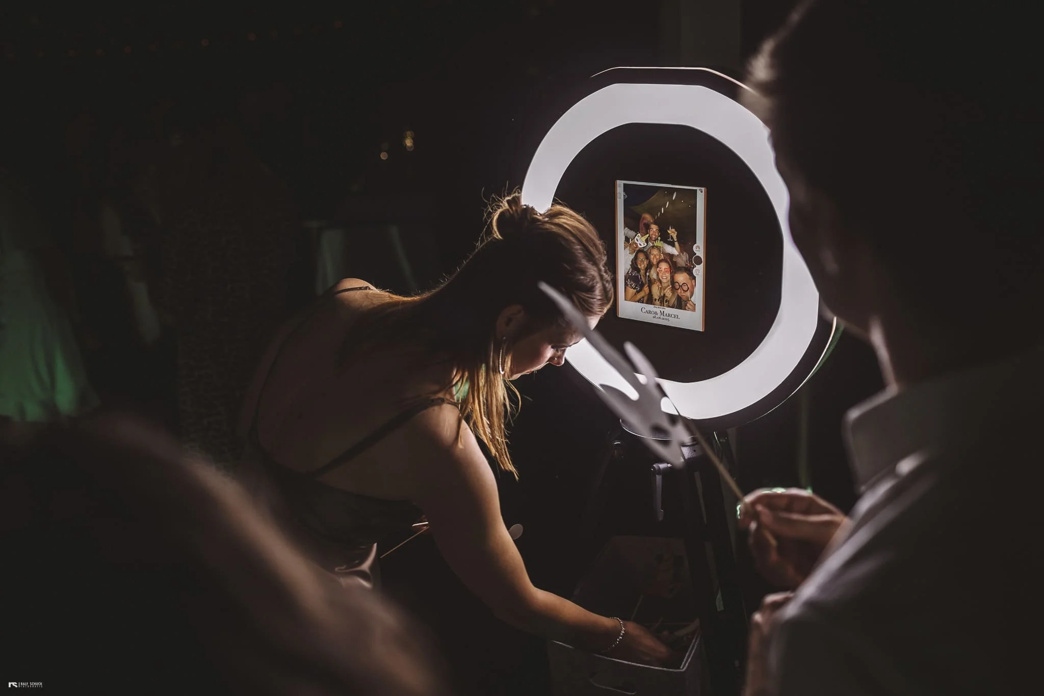 Gruppen von Menschen, die in einer Fotokabine Fotos machen, man sieht eine Frau, die sich gerade vor der Kamera bückt, und eine Person, die etwas in der Hand hält, während die Fotokabine ein Licht rund um das Kamera-Display hat.