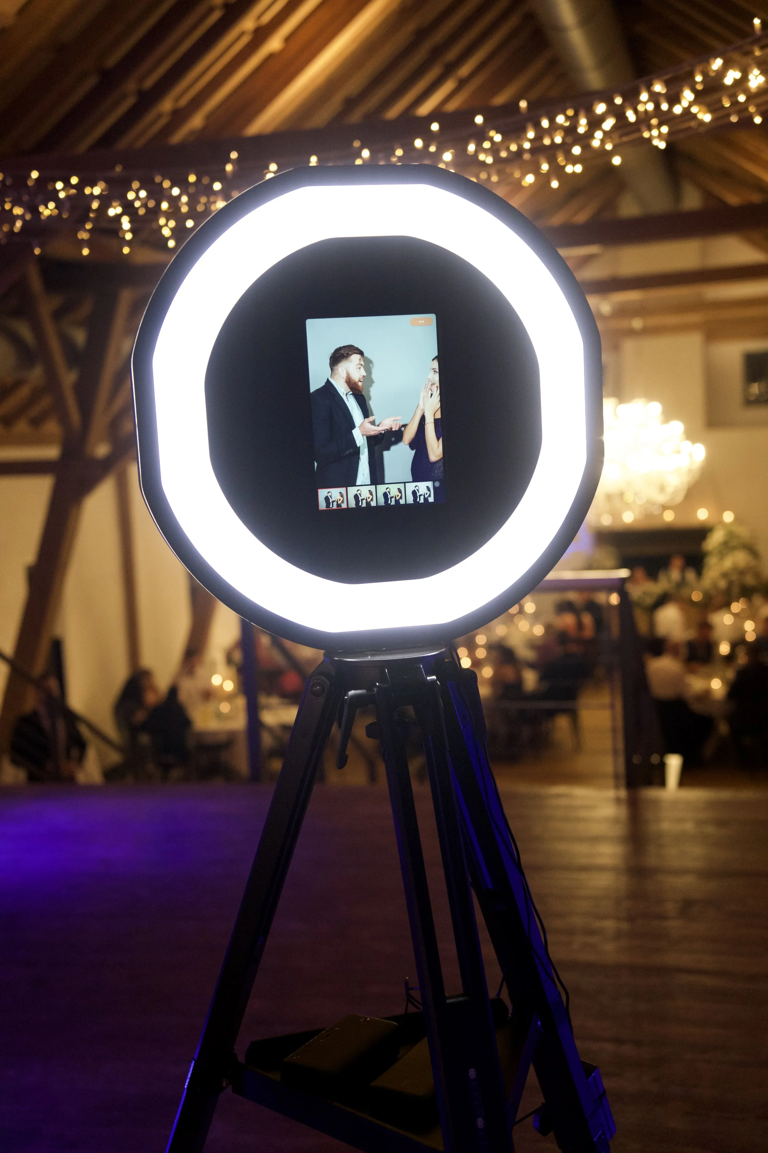 Fotobox mit Ringlicht und einem Foto im Display, das ein Mann und eine Frau beim Lachen und Fotografieren zeigt, im Hintergrund eine festliche Dekoration mit Lichterketten, Menschen und einem Saal.