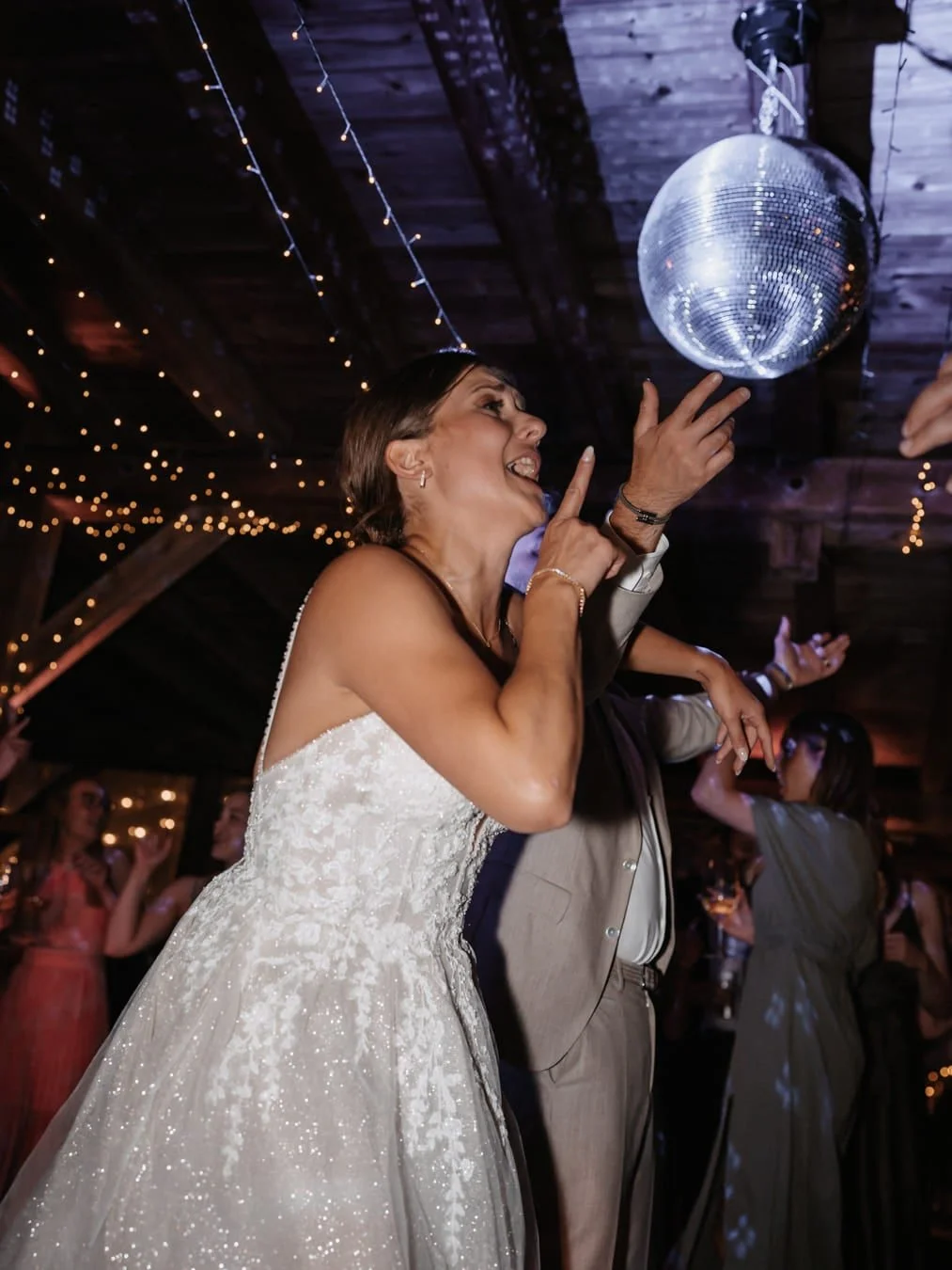 Eine lachende Frau in einem weißen Brautkleid tanzt bei einer Feier unter einer Discokugel, umgeben von anderen Gästen mit Lichtern an der Decke.