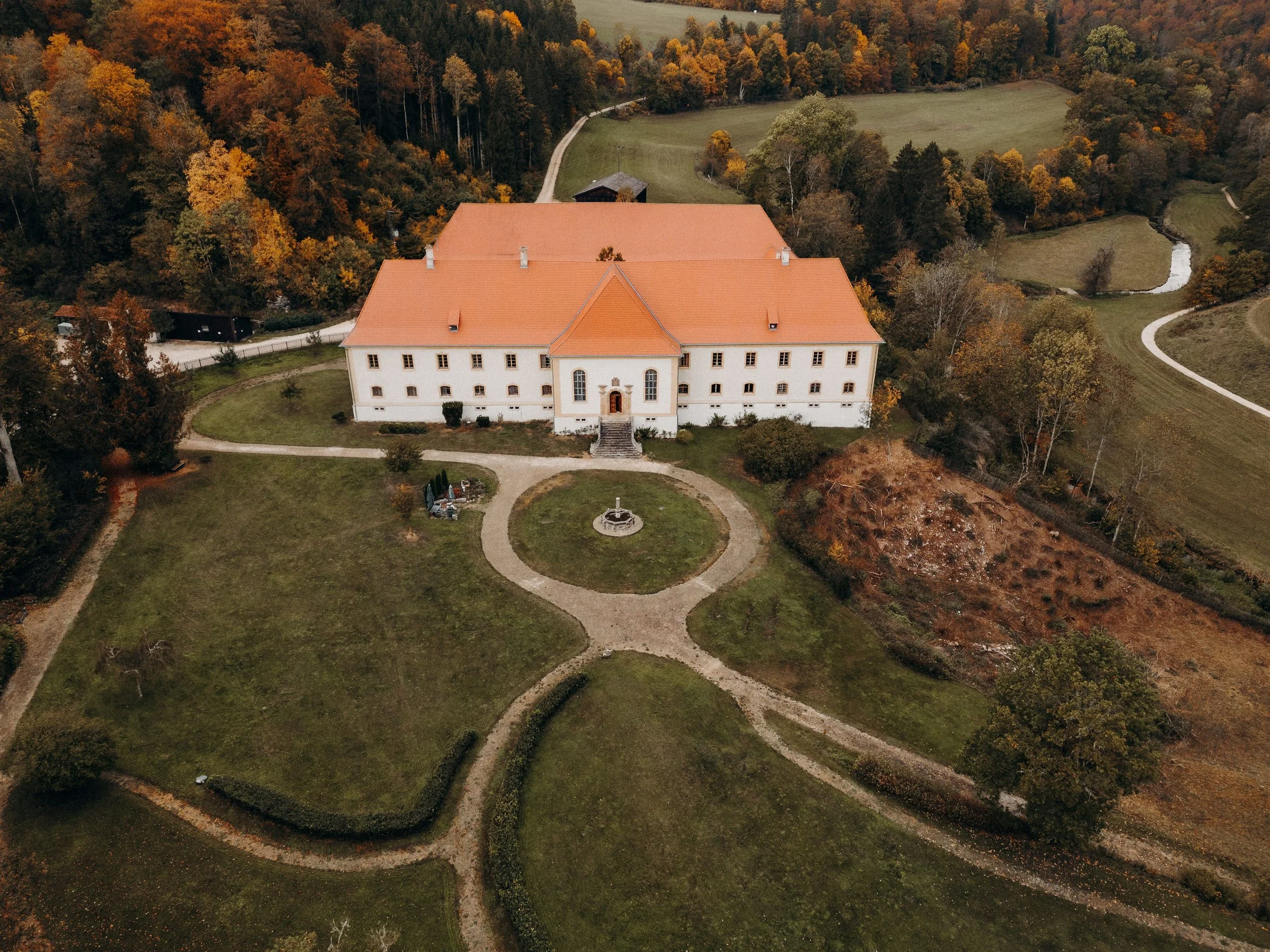 Ein weißes Gebäude mit rotem Dach inmitten eines gepflegten Gartens, umgeben von Bäumen im Herbst, mit Wegen und einem Brunnen im Vordergrund.