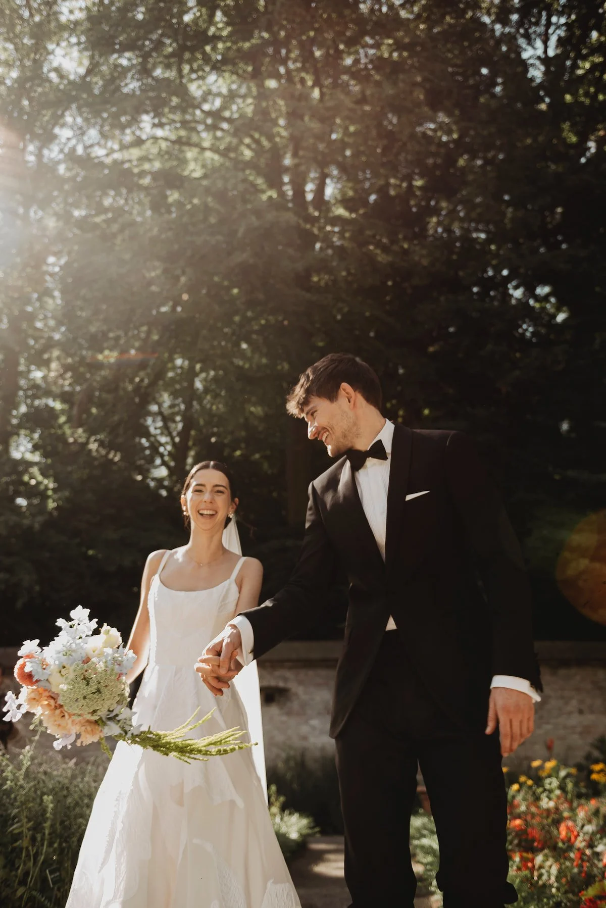 Ein lachendes Brautpaar im Garten bei Sonnenlicht, die Braut trägt ein weißes Kleid mit Trägern und hält einen Blumenstrauß, der Bräutigam trägt einen schwarzen Anzug mit Fliege, sie halten sich bei einer Hochzeitshand in Hand, im Hintergrund sind Bäume und bunte Blumen sichtbar.