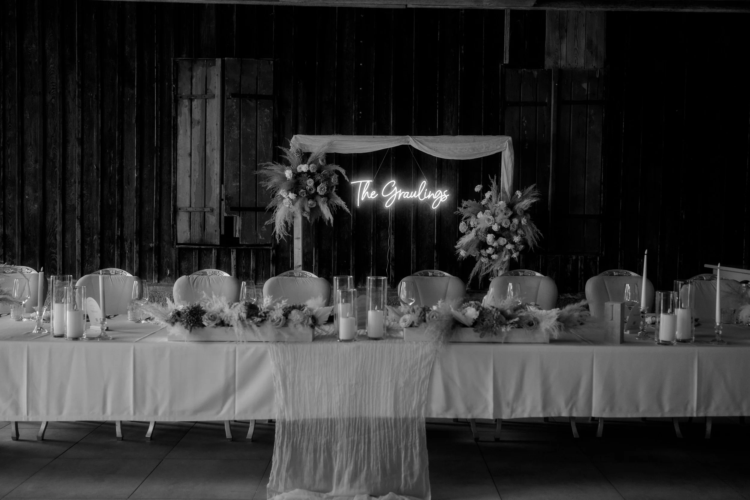 Ein festlich dekorierter Tisch mit Kerzen, Blumen und einem Neon-Schild mit der Aufschrift "The Graulings" vor einer dunklen Holzvertäfelung.