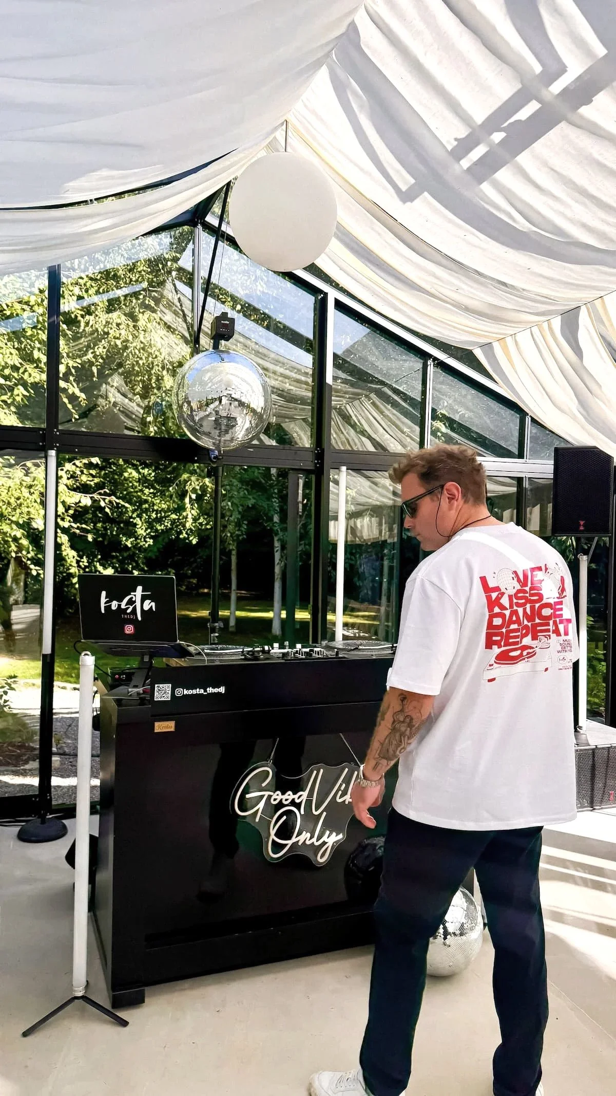 Ein junger Mann mit Tattoo, Sonnenbrille und einem weißen T-Shirt, das auf der Rückseite die Worte »Love Kiss Dance Repeat« in Rot und Schwarz hat, steht vor einem DJ-Pult im Freien. Im Hintergrund ist ein Glasgebäude mit großen Fenstern, durch die Bäume und die Natur sichtbar sind. Es gibt eine Diskokugel, einen weißen runden Lampenschirm und eine Tafel mit dem Wort »Good Vibes Only« auf dem DJ-Pult.