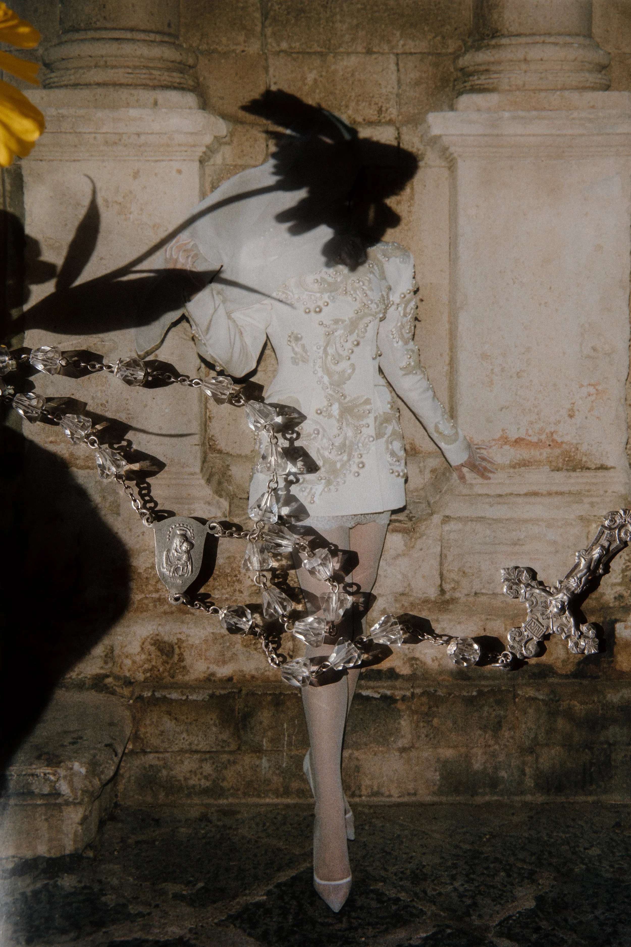 Person in an ornate white outfit with pearls and lace, wearing a black hat, standing against a stone wall. There is a large decorative chain in the foreground.