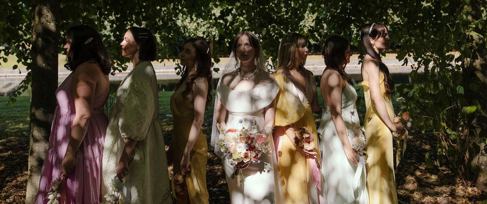 A group of seven women standing outdoors in a line, wearing dresses in various pastel colors such as pink, yellow, and white. The central figure wears a white bridal gown holding a bouquet. They are under the shade of trees, with dappled sunlight creating patterns on their dresses and the ground.
