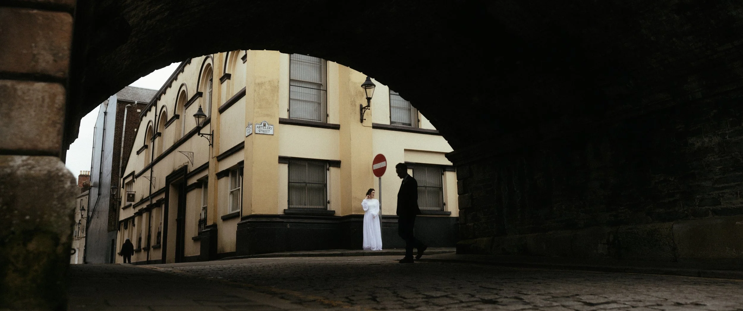View through a dark archway of a street scene with a person in white standing near a corner building, a street sign, and a person walking by. The building has vintage-style architecture with arched windows and lamps.