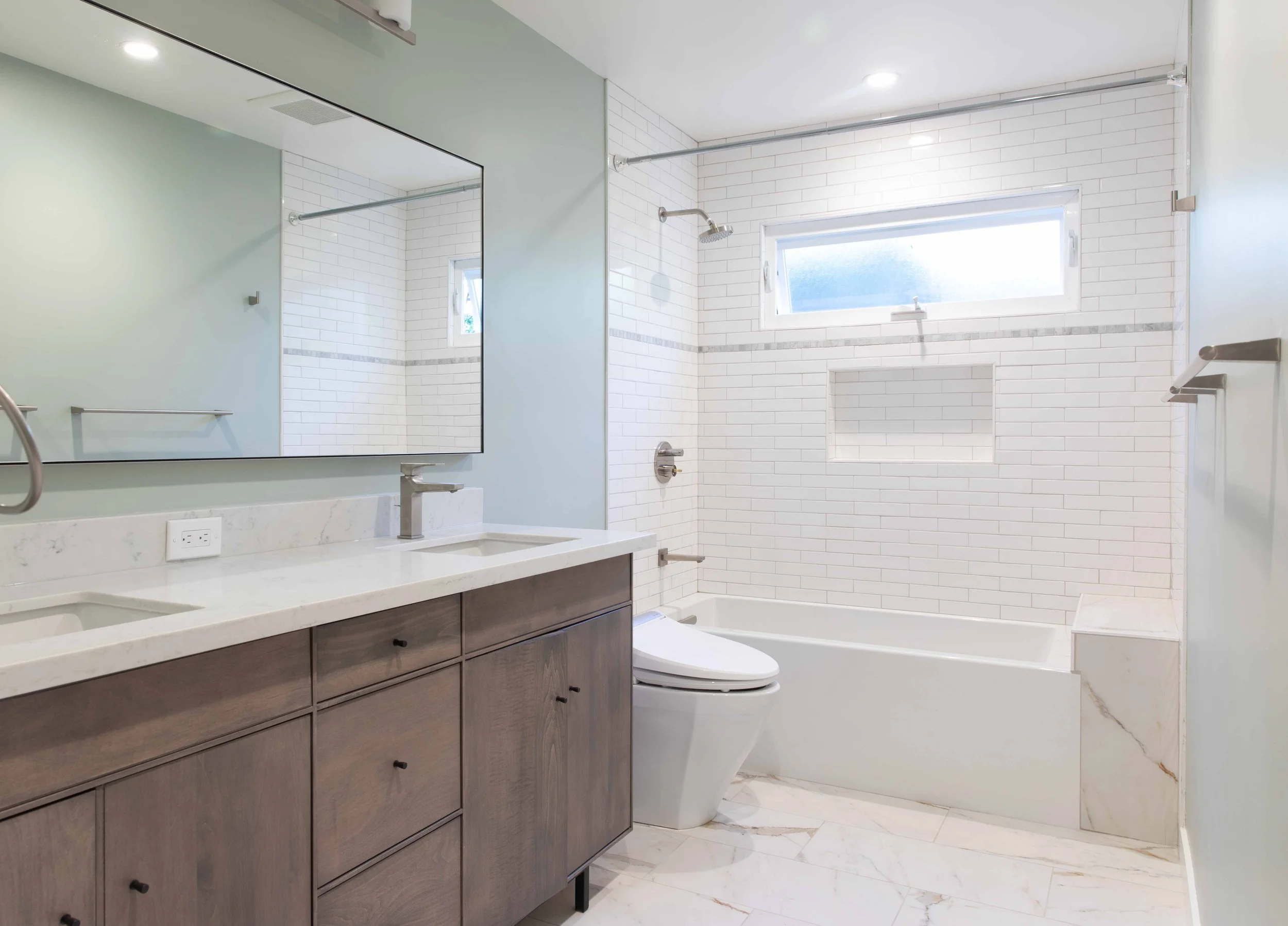 Modern bathroom with double vanity, large mirror, white countertops, gray wood cabinets, toilet, bathtub with white subway tile walls, built-in niche, small window, and marble tile flooring.