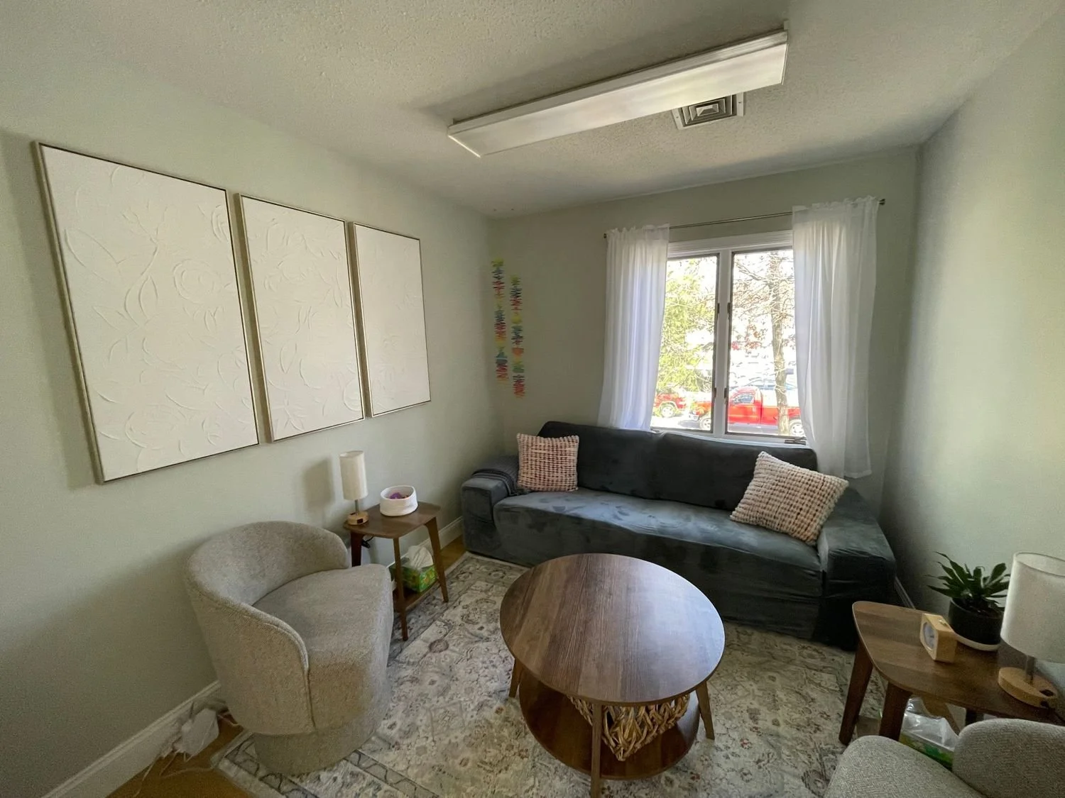 Living room with light green walls, a black couch near a window with white curtains, a beige upholstered armchair, a wooden coffee table, a side table with a lamp and small decorative items, a potted plant, framed white textured art on the wall, and a patterned area rug.