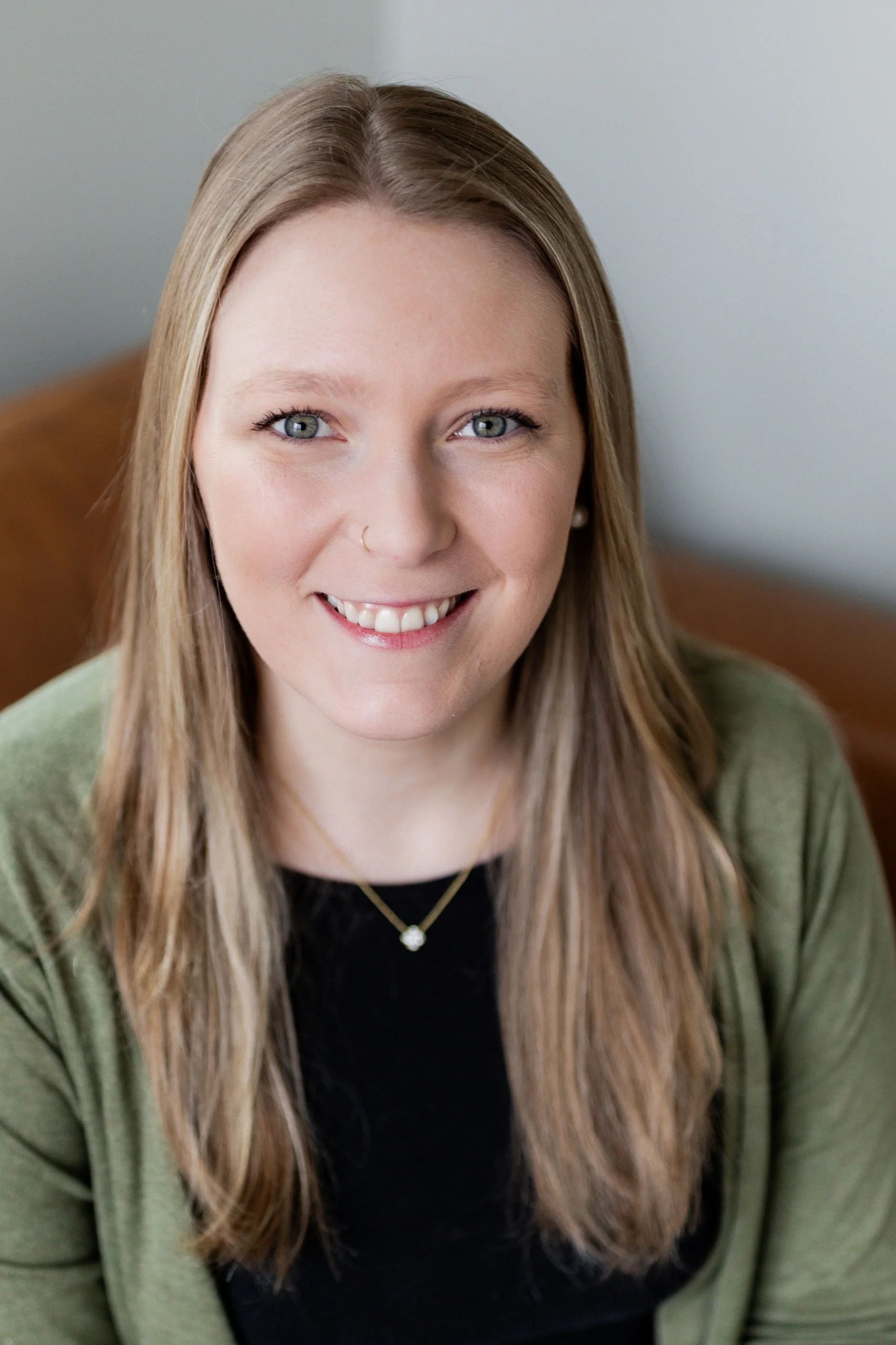 Close-up of a young woman with long light brown hair, smiling, wearing a black top and a green cardigan, sitting indoors against a plain wall.