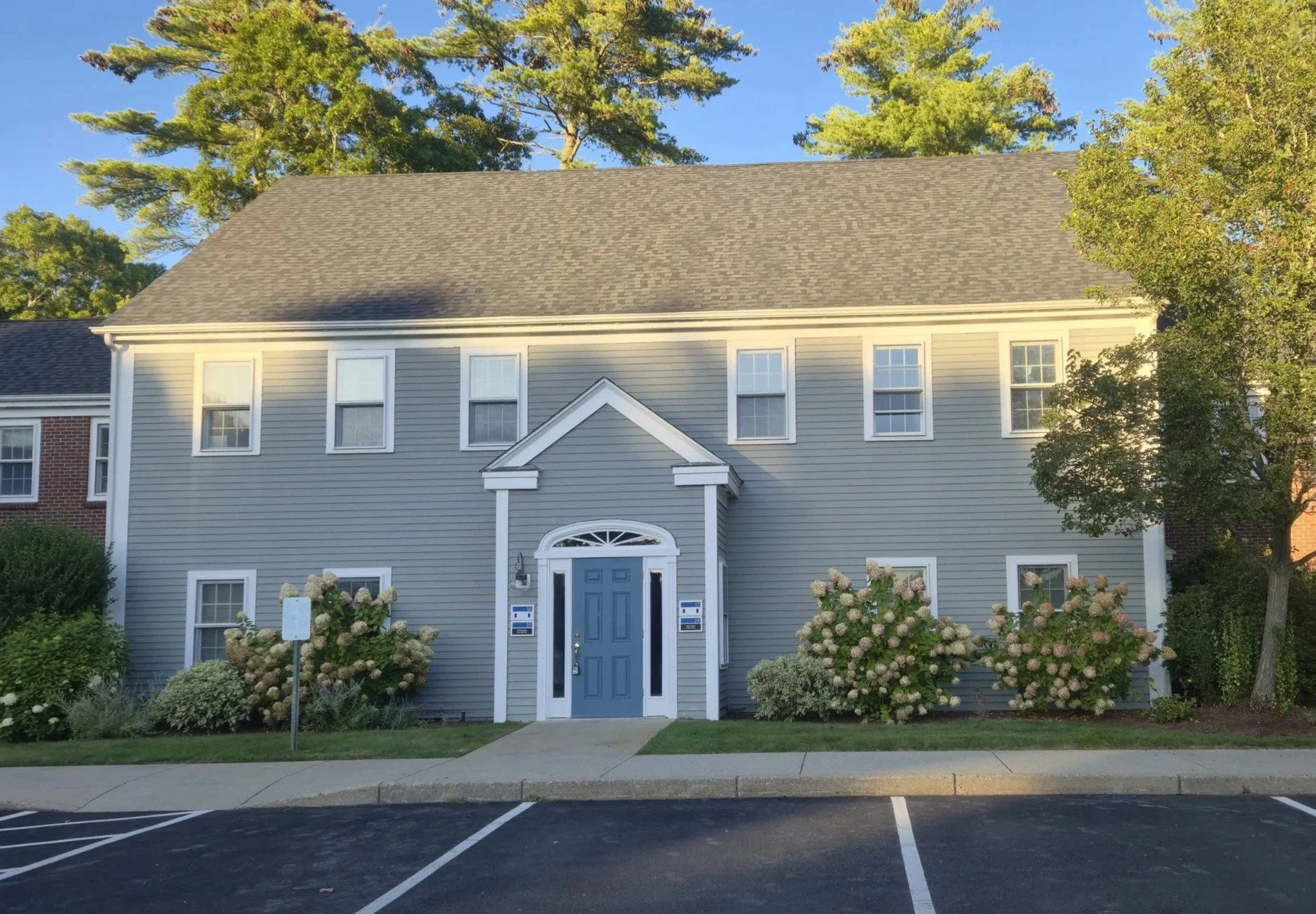 Front view of a three-story residential building with gray siding, white trim, and a blue front door, surrounded by bushes and flowers, with parking spaces and a sidewalk in the foreground.