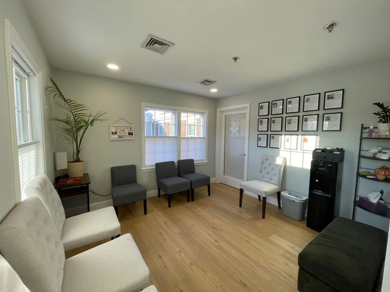 Interior of a waiting room with multiple chairs, a water cooler, framed documents on the wall, a potted plant, and large windows allowing natural light.