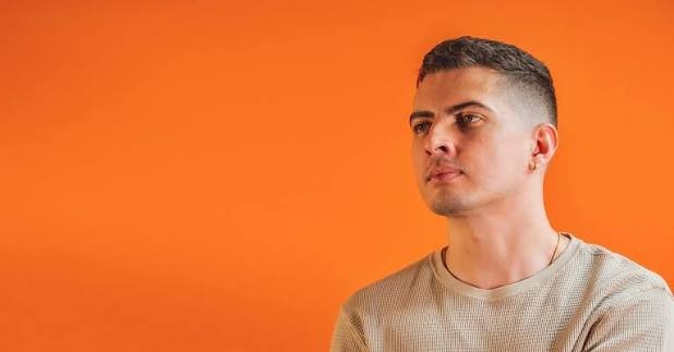 DJ OMNAM - A young man with short dark hair and a serious expression, wearing a beige shirt, standing against an orange background.