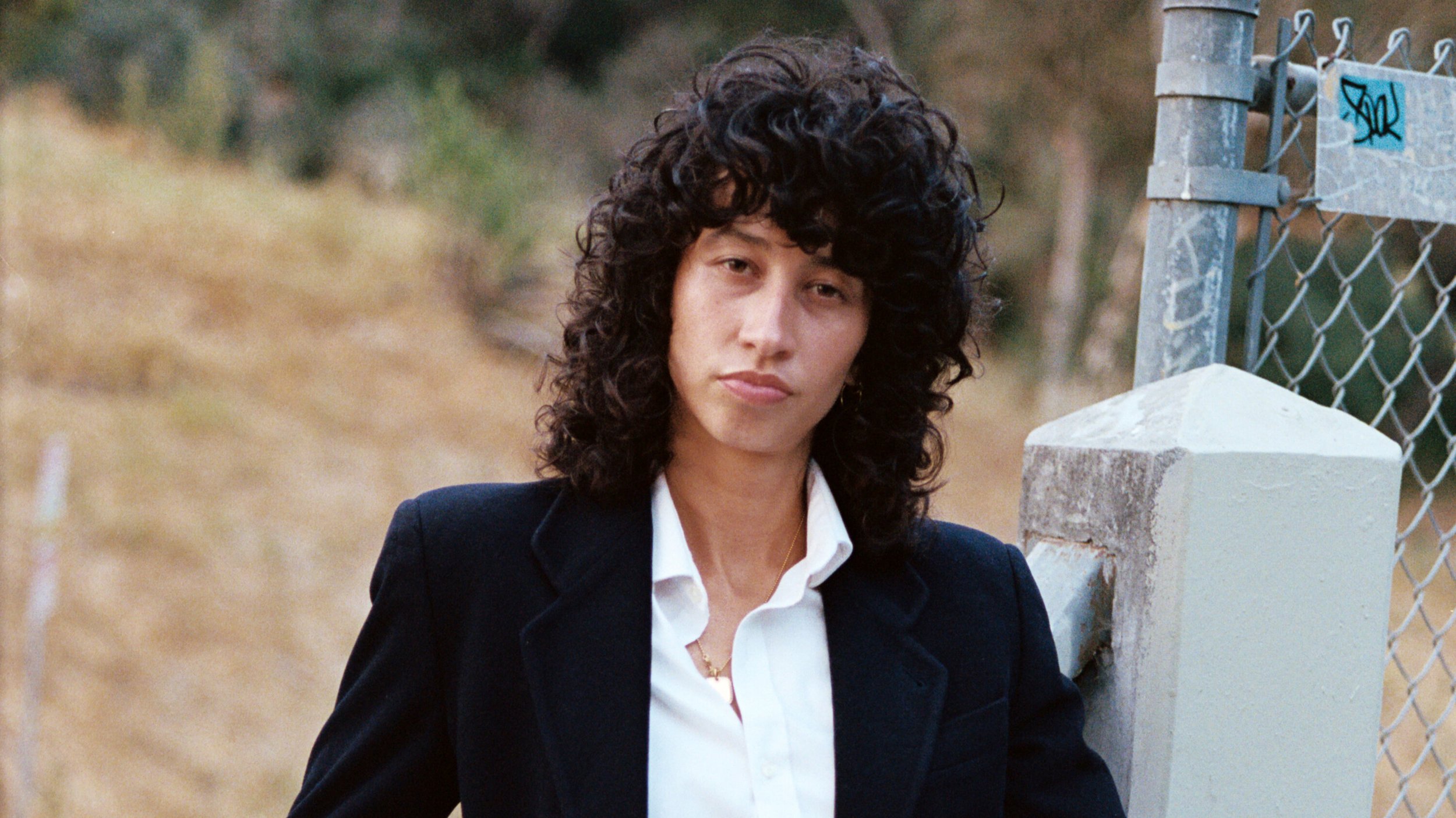 Towa Bird. A woman with dark curly hair wearing a black blazer and white shirt standing beside a chain-link fence with an outdoor background.