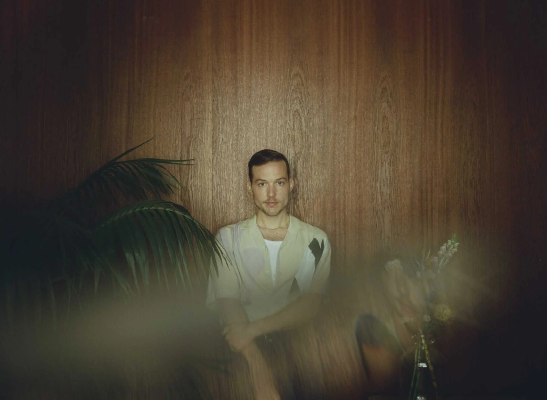 Ben Böhmer. A young man with short dark hair and a light-colored shirt standing in front of a wooden wall with plants partially obscuring the lower part of the image.