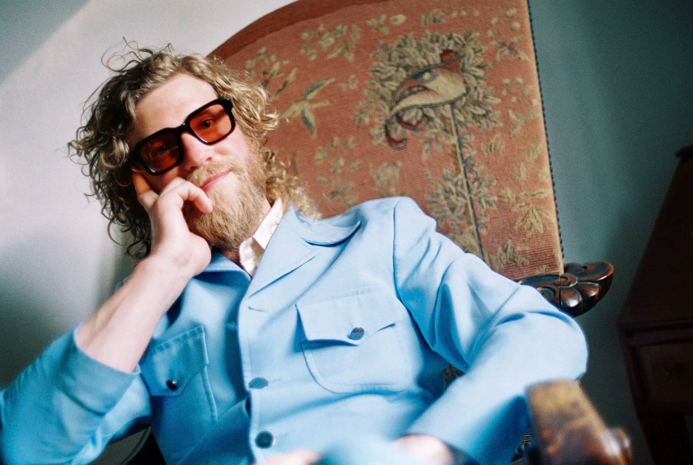 Soul R&B Singer, Allen Stone, with curly hair, beard, sunglasses, and a light blue shirt, sitting in a vintage chair with a patterned tapestry in the background.
