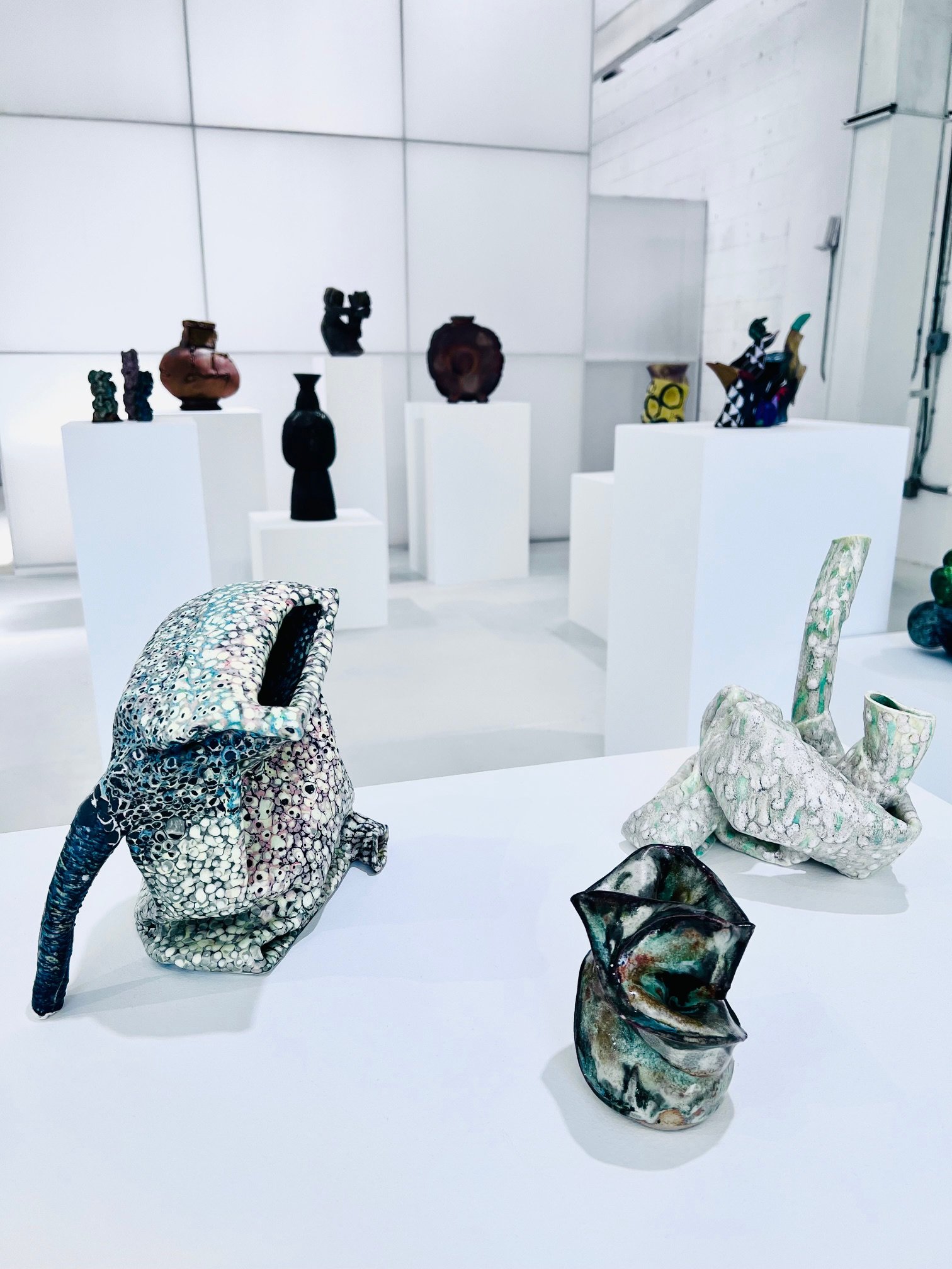 Colorful ceramic sculptures of various abstract shapes and figures displayed on white pedestals and table in an art gallery with white walls.