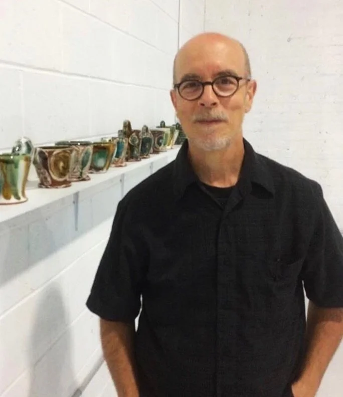 A man with glasses and a bald head standing in front of a white brick wall with ceramic cups on a shelf behind him.