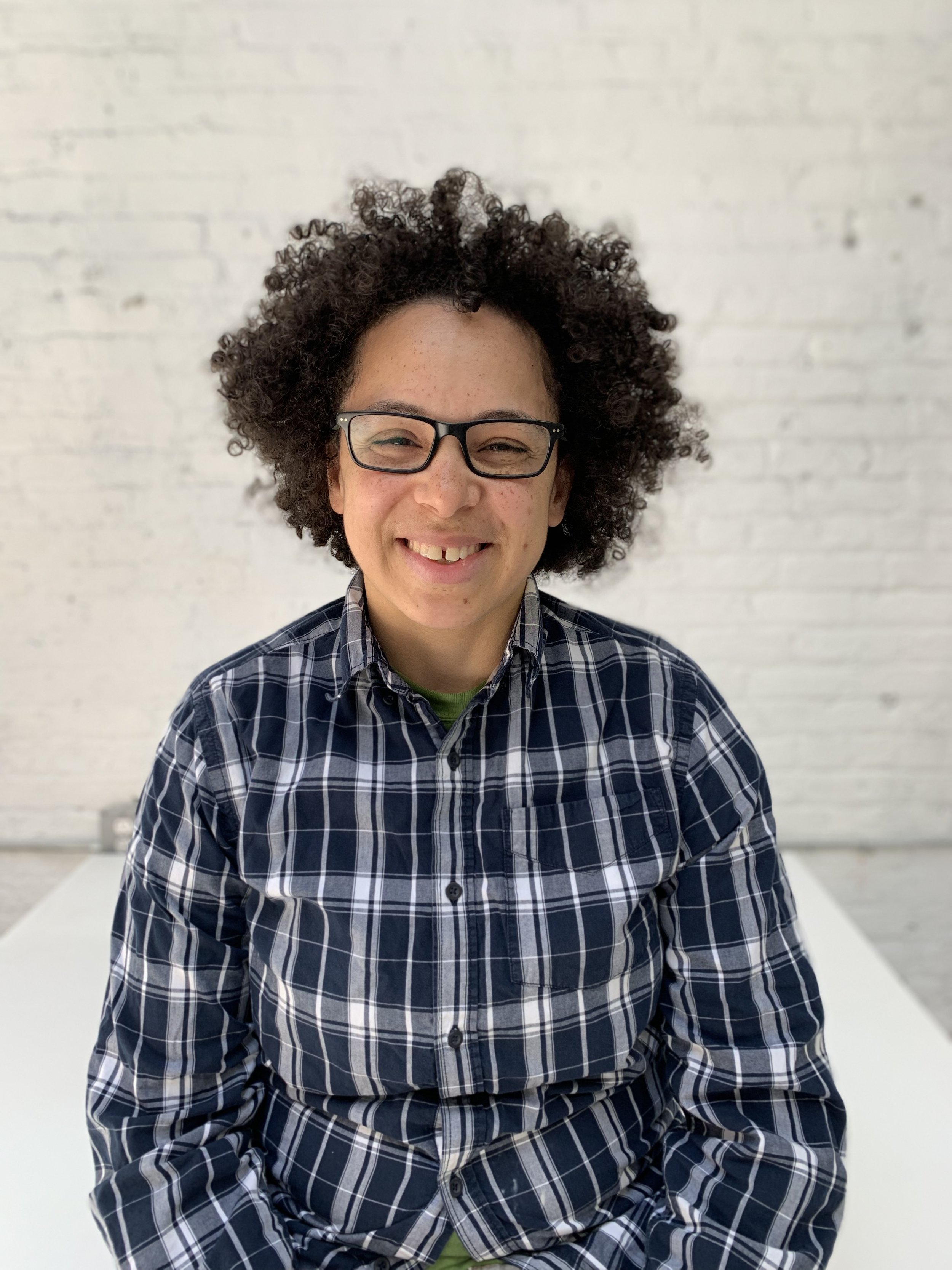 A person with curly hair, glasses, and a plaid shirt smiling in front of a white brick wall.