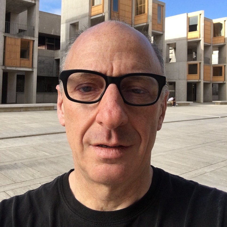 Close-up of a middle-aged man with glasses in front of modern apartment buildings.