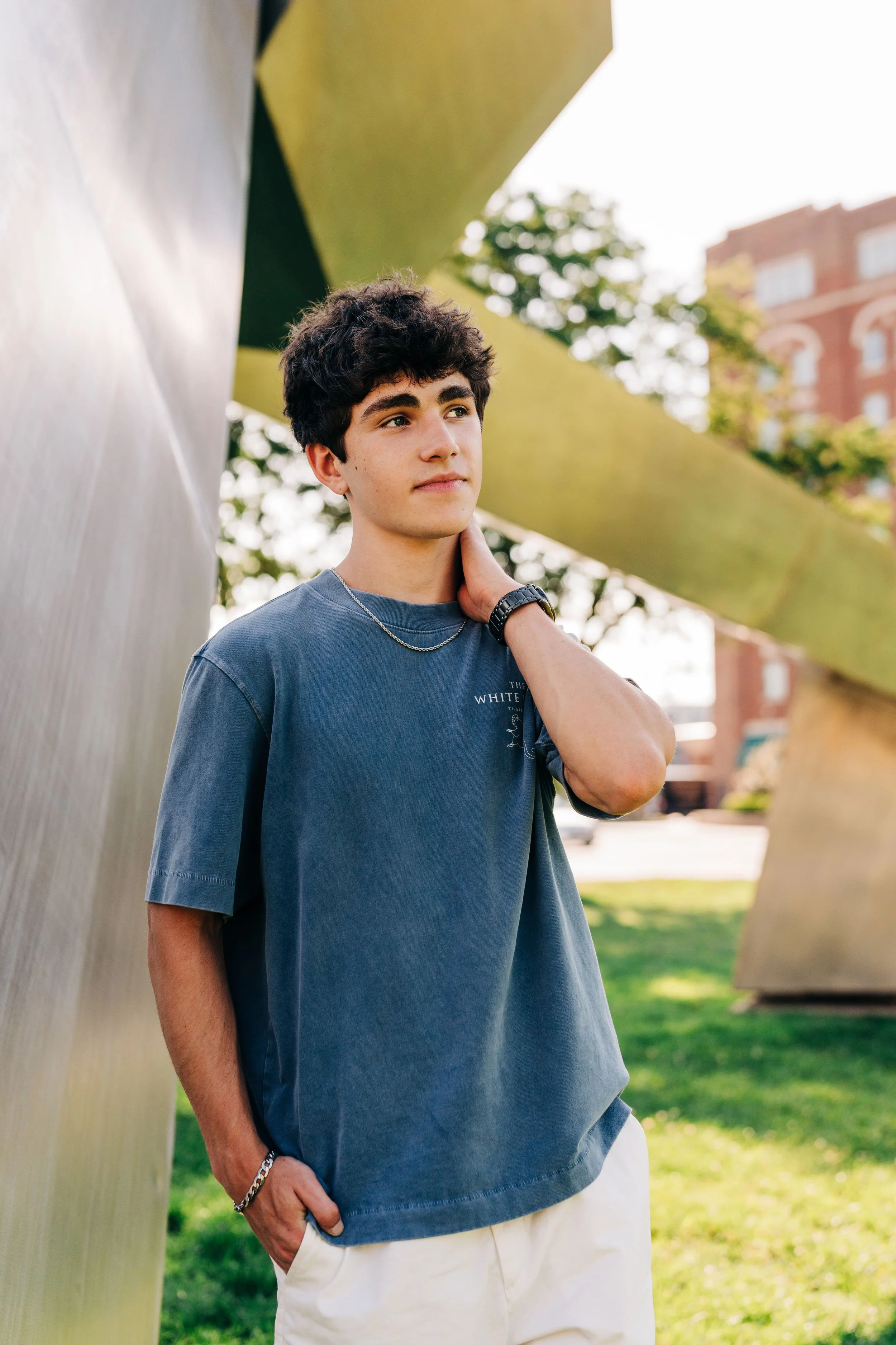 A young man with dark, curly hair, wearing a blue T-shirt, light-colored pants, a silver chain, and a black watch, stands outdoors near an abstract metallic sculpture. He is touching his neck with his right hand and has his left hand in his pocket, l
