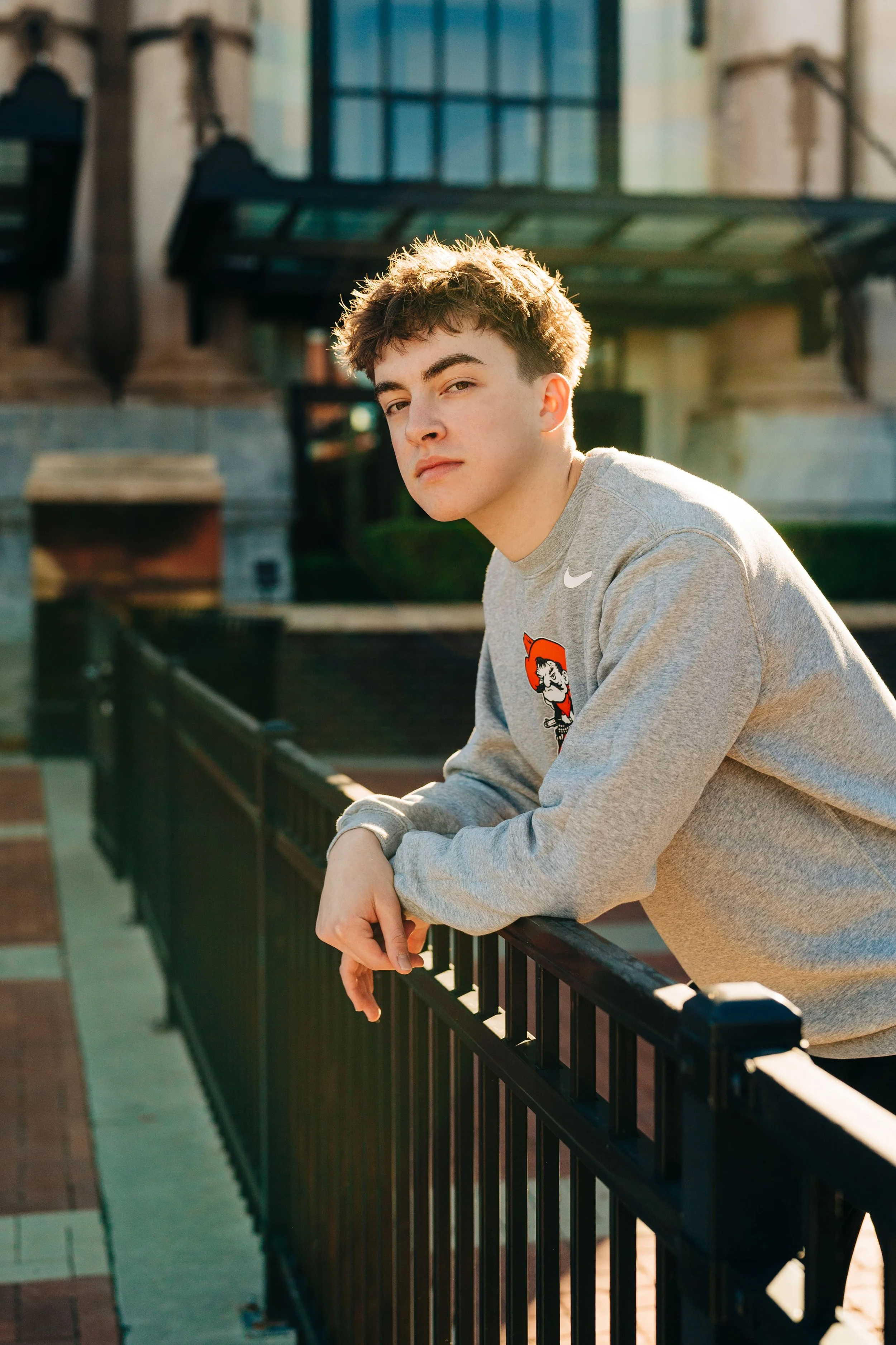 Young man leaning on a black metal railing outdoors, wearing a grey sweatshirt with a cartoon character patch and the Nike logo, with a building in the background during sunlight.