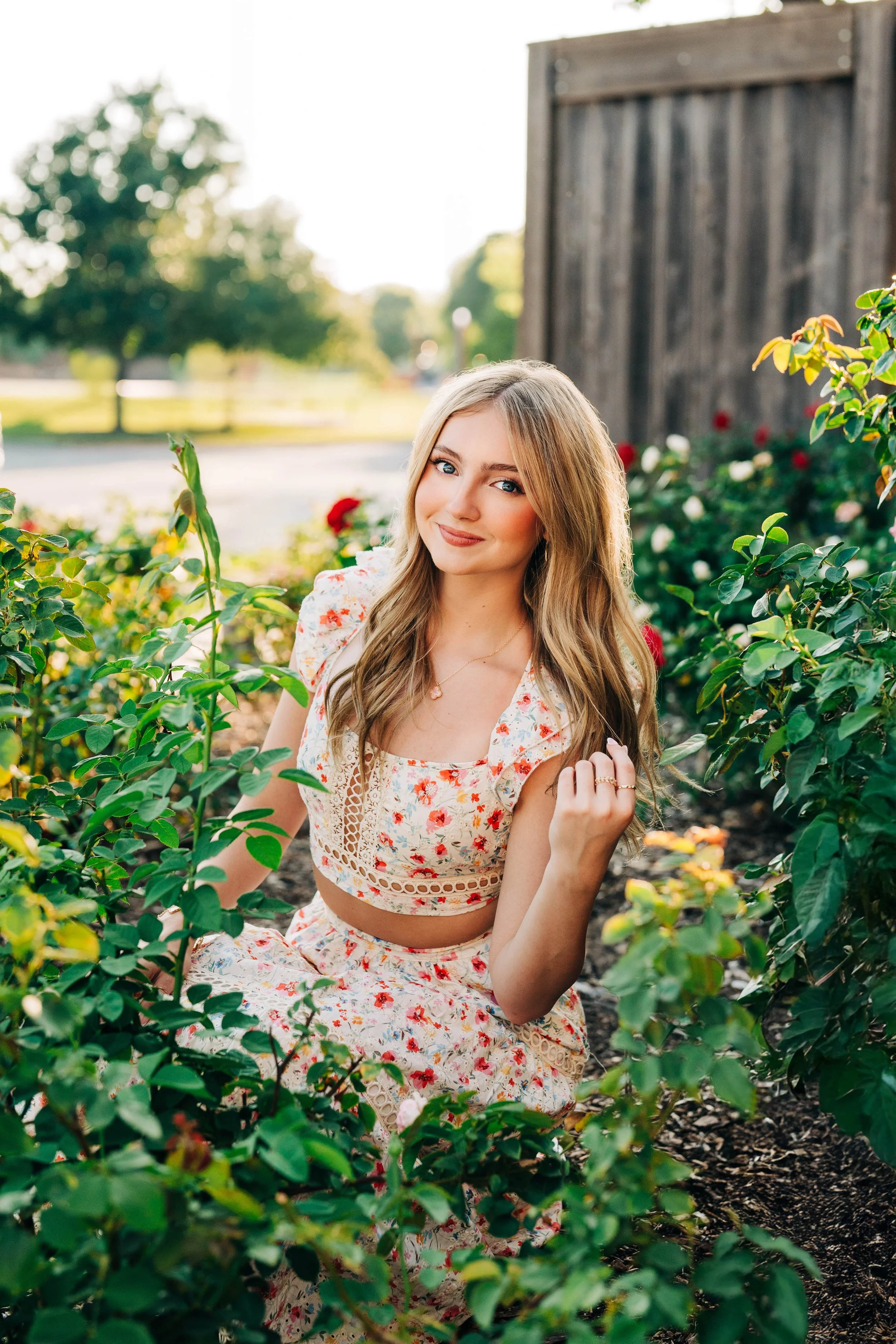 A young woman with long blonde hair and blue eyes sitting among green bushes and flowers outdoors, smiling at the camera. She is wearing a floral crop top and matching skirt, with a small necklace and rings.
