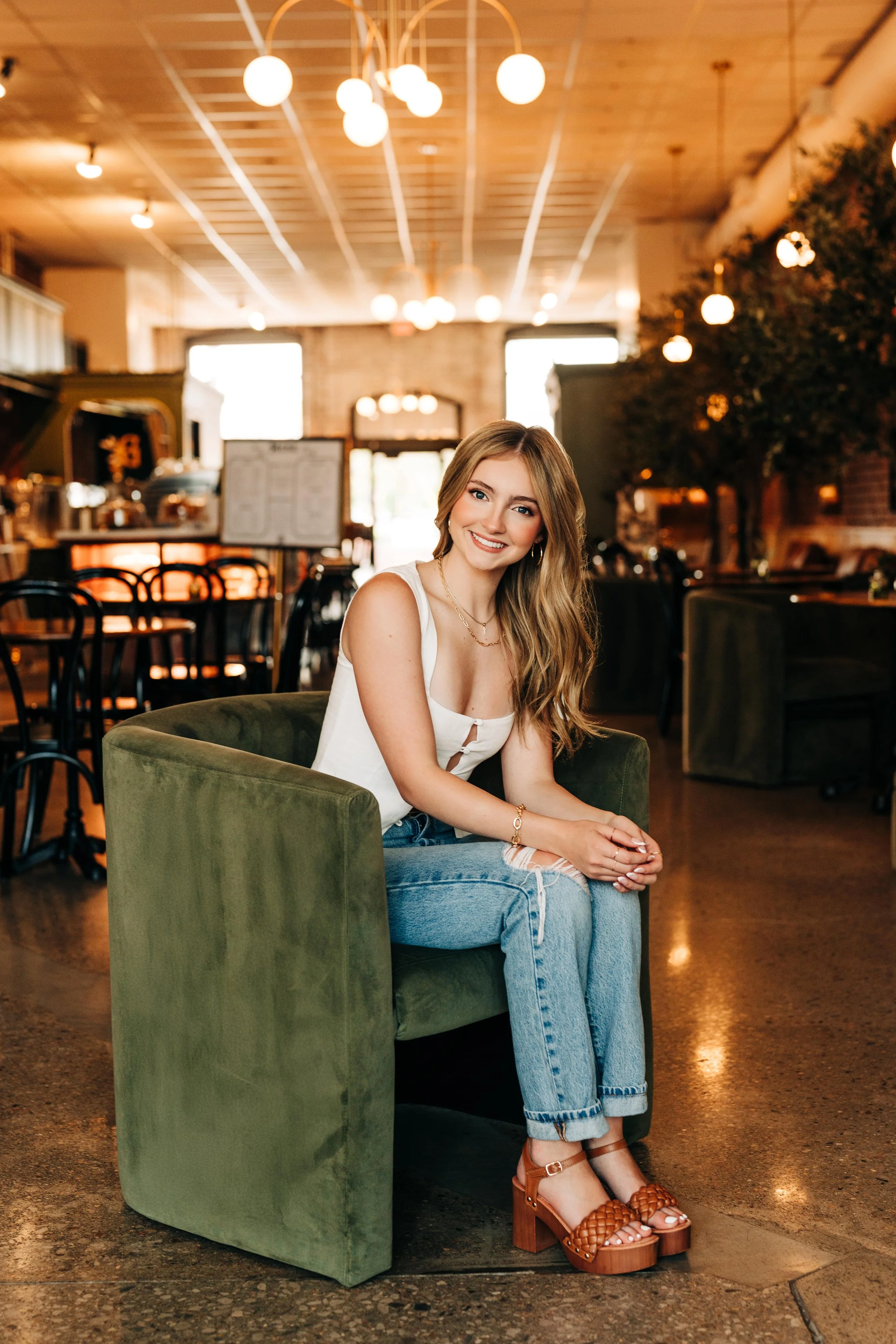 A young woman with long wavy hair, wearing a white sleeveless top, ripped jeans, and brown platform sandals, sitting on a green armchair inside a warmly lit cafe or restaurant and smiling at the camera.