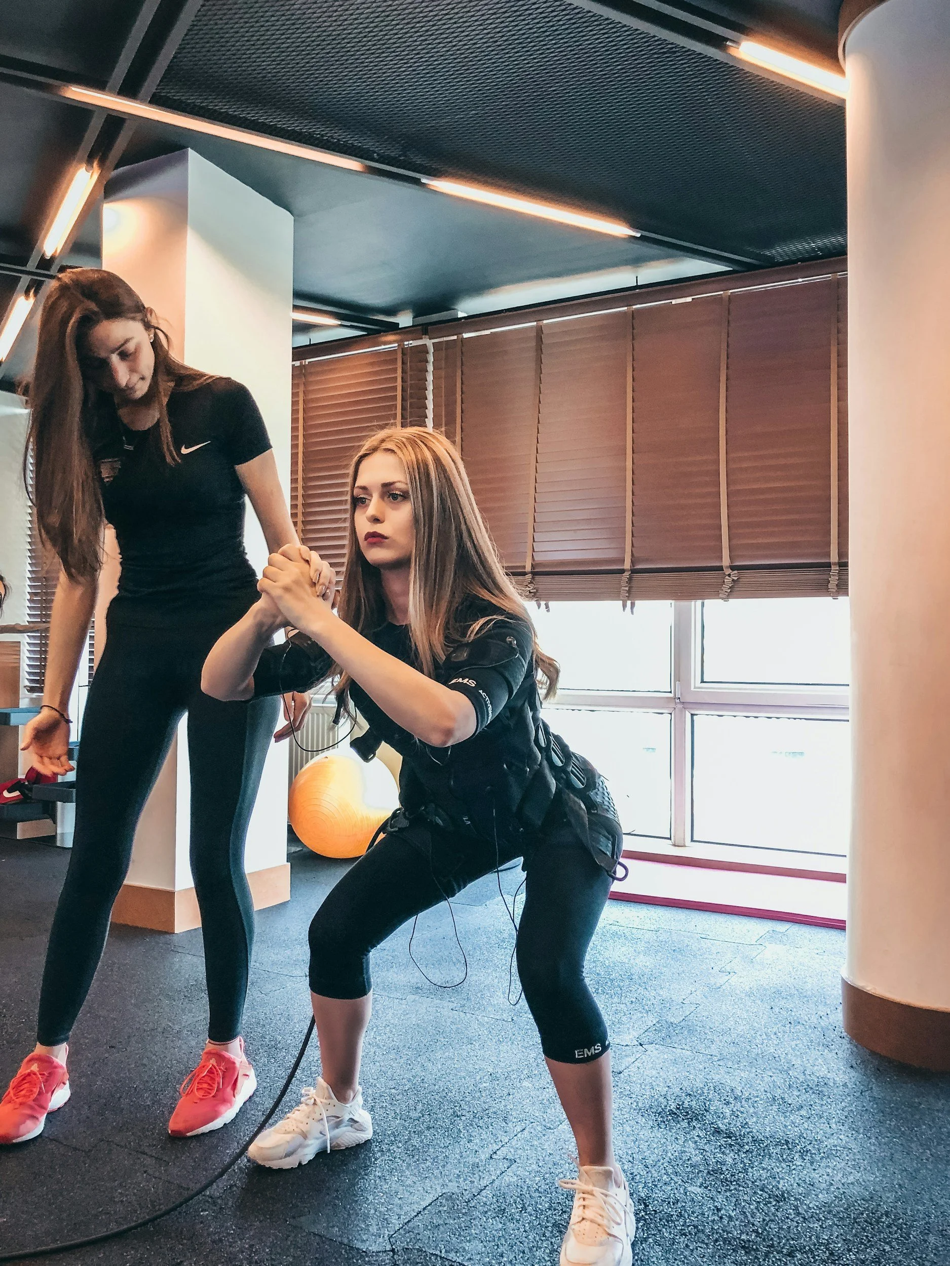 A woman is performing a squat exercise with a trainer providing assistance, inside a gym with wood blinds and large windows.